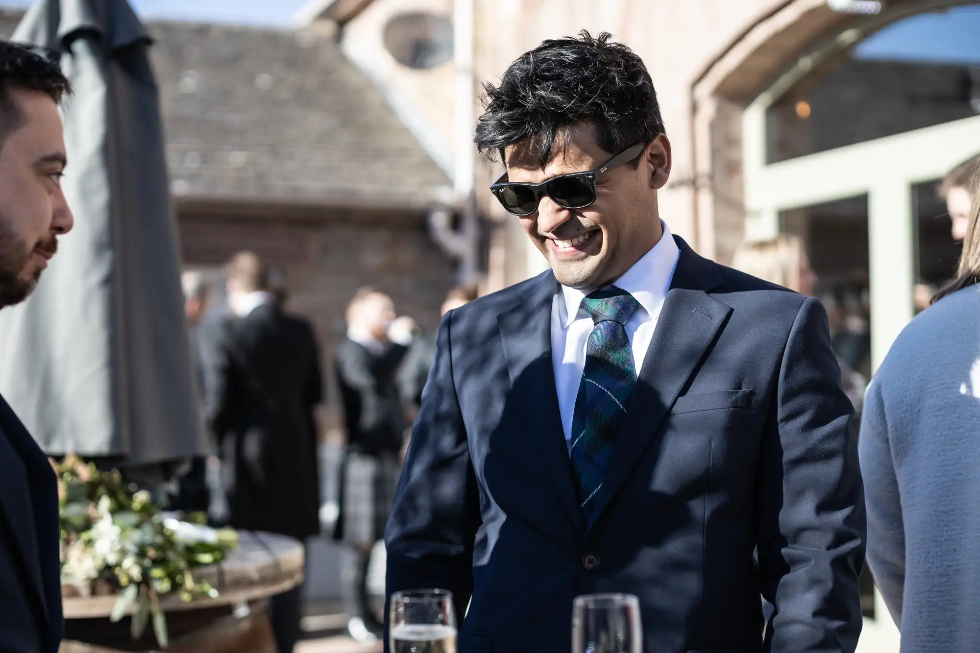 Man in a suit and sunglasses smiling at an outdoor event, holding a drink. Other people can be seen socializing in the background.