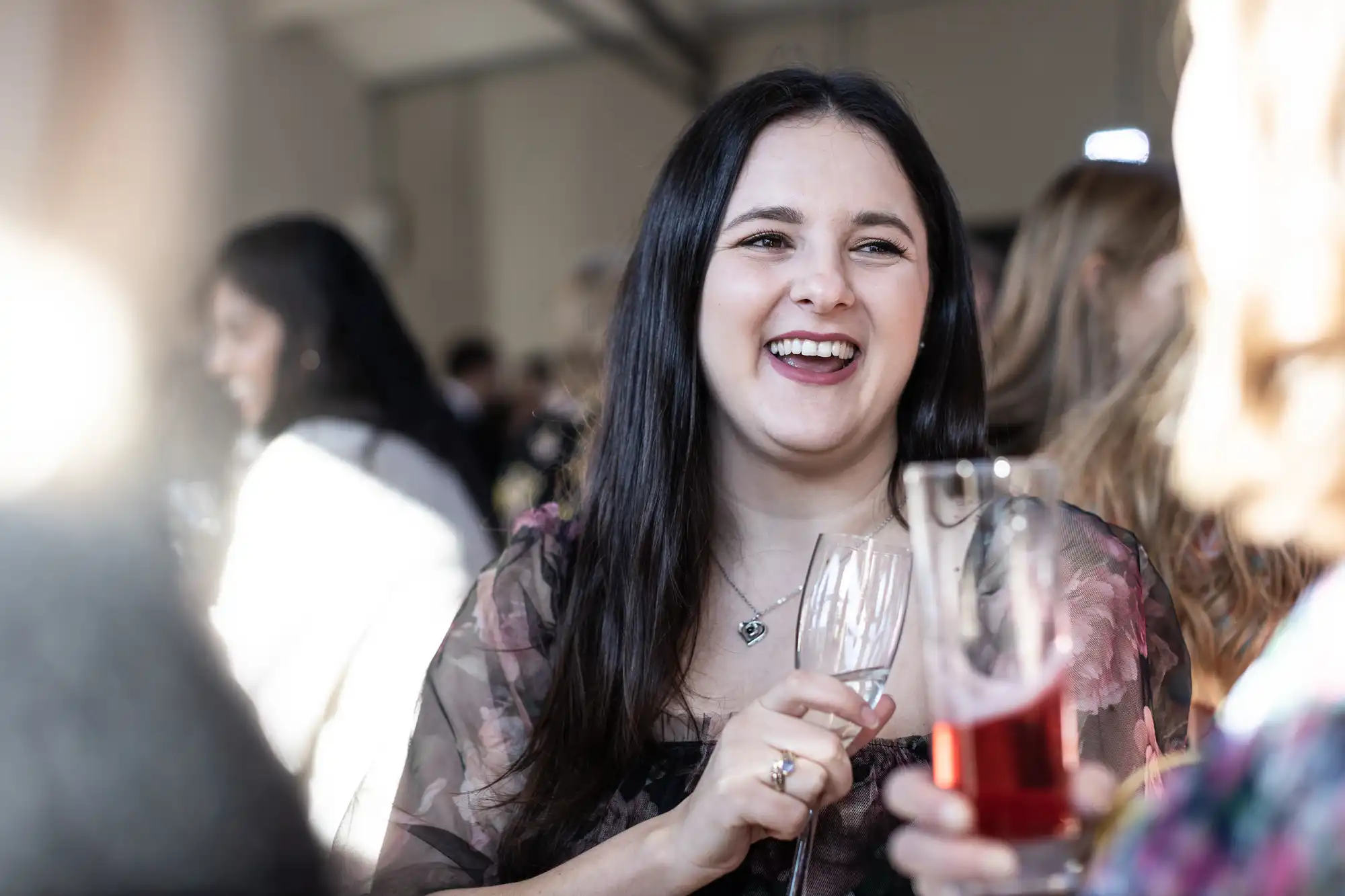 A woman with long dark hair smiles while holding a glass of wine at a social gathering.