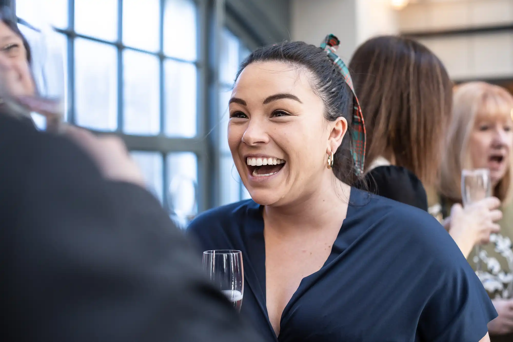 A woman in a blue shirt smiles and holds a drink while conversing with others at an indoor social gathering.