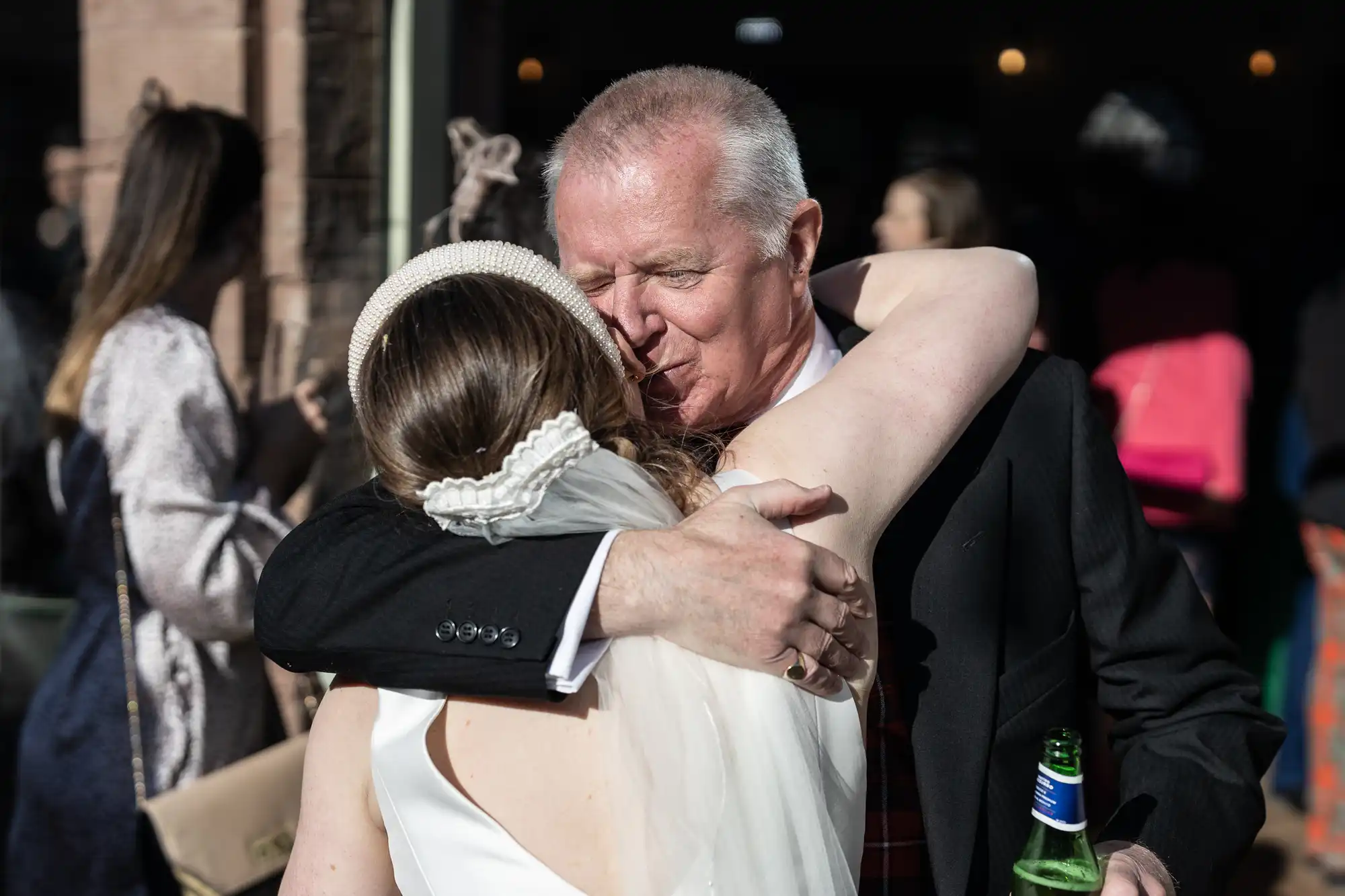 A person in a wedding dress embraces an older man holding a beer bottle. They are outdoors among a group of people.