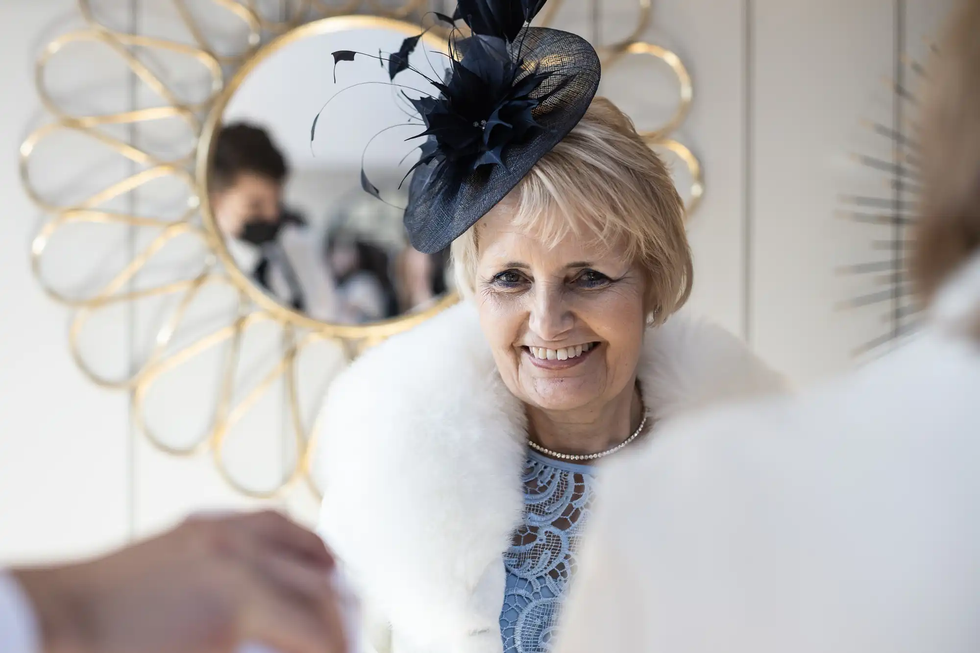 A smiling woman wearing a black hat with decorative feathers and a white fur collar stands in front of a decorative mirror.