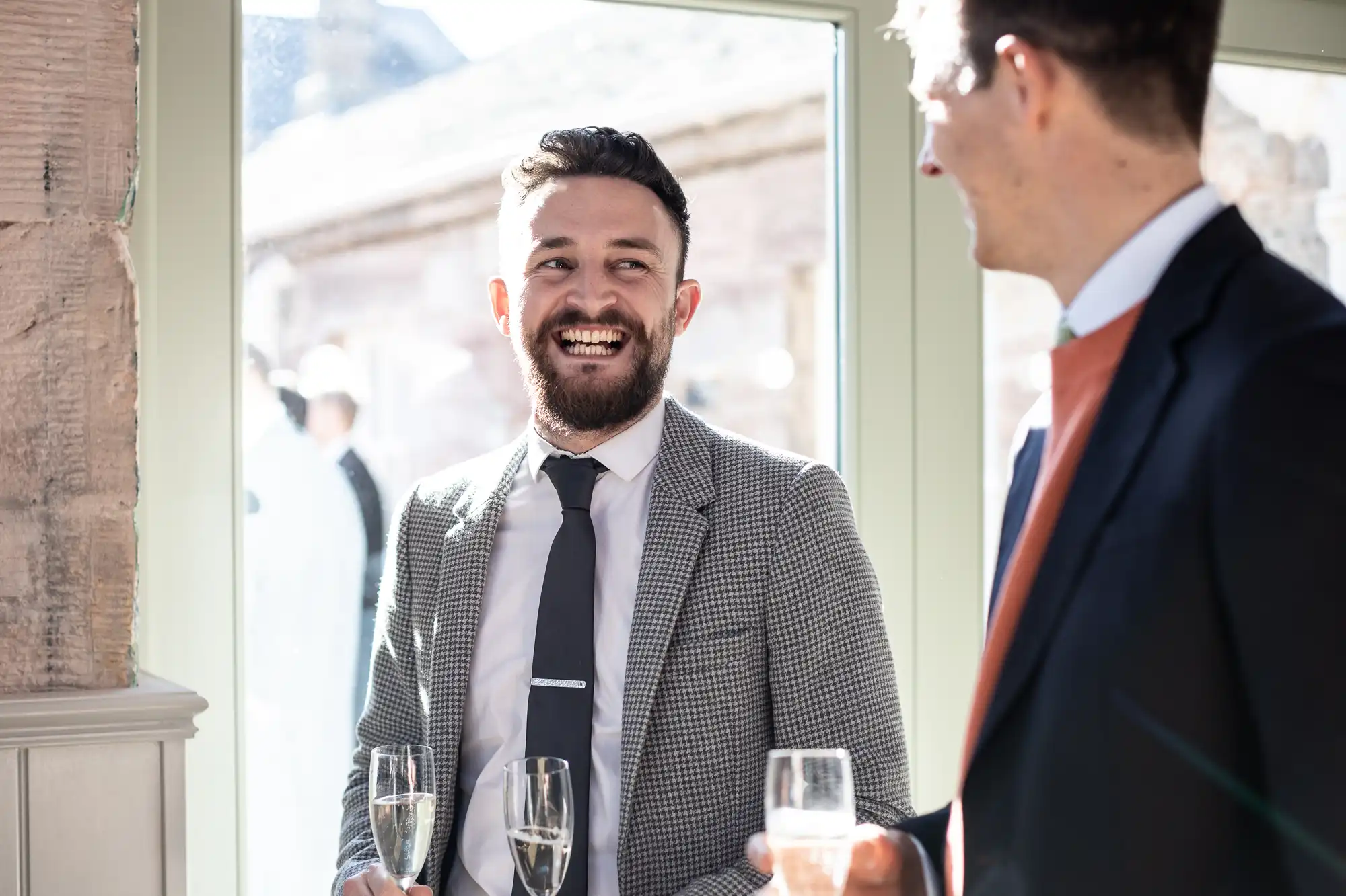 Two men, dressed formally in suits, are conversing and smiling while holding glasses of champagne near a window.