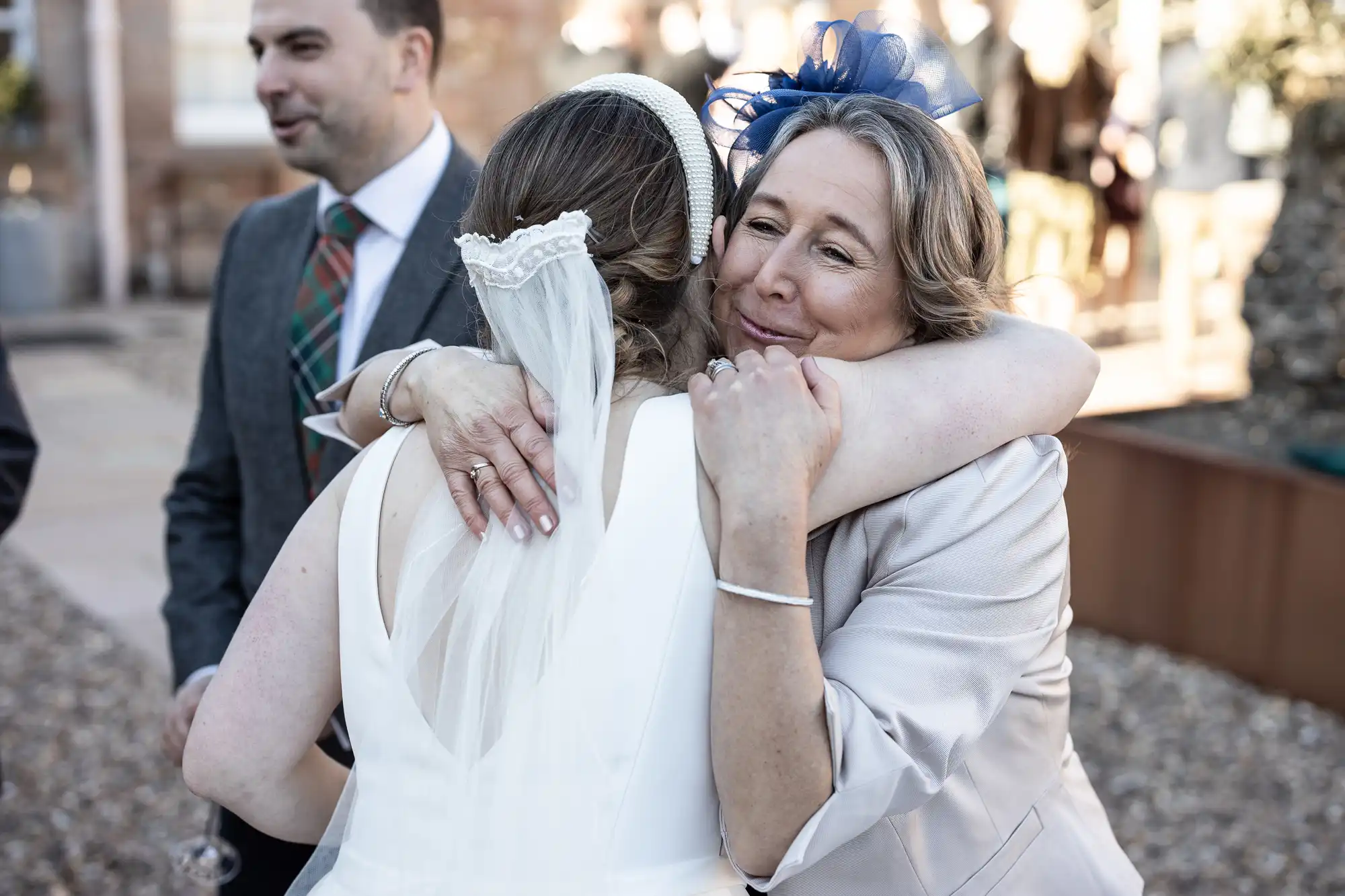 Two women embrace at an outdoor event, one wearing a white dress and veil, and the other a cream outfit with a blue fascinator. A man in a suit and patterned tie stands in the background.