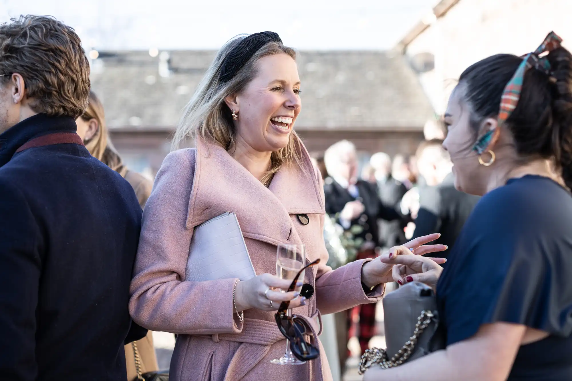 Two women engage in a lively conversation outdoors; one wearing a pink coat and holding a wine glass and clutch, the other in a dark outfit with a hair accessory.