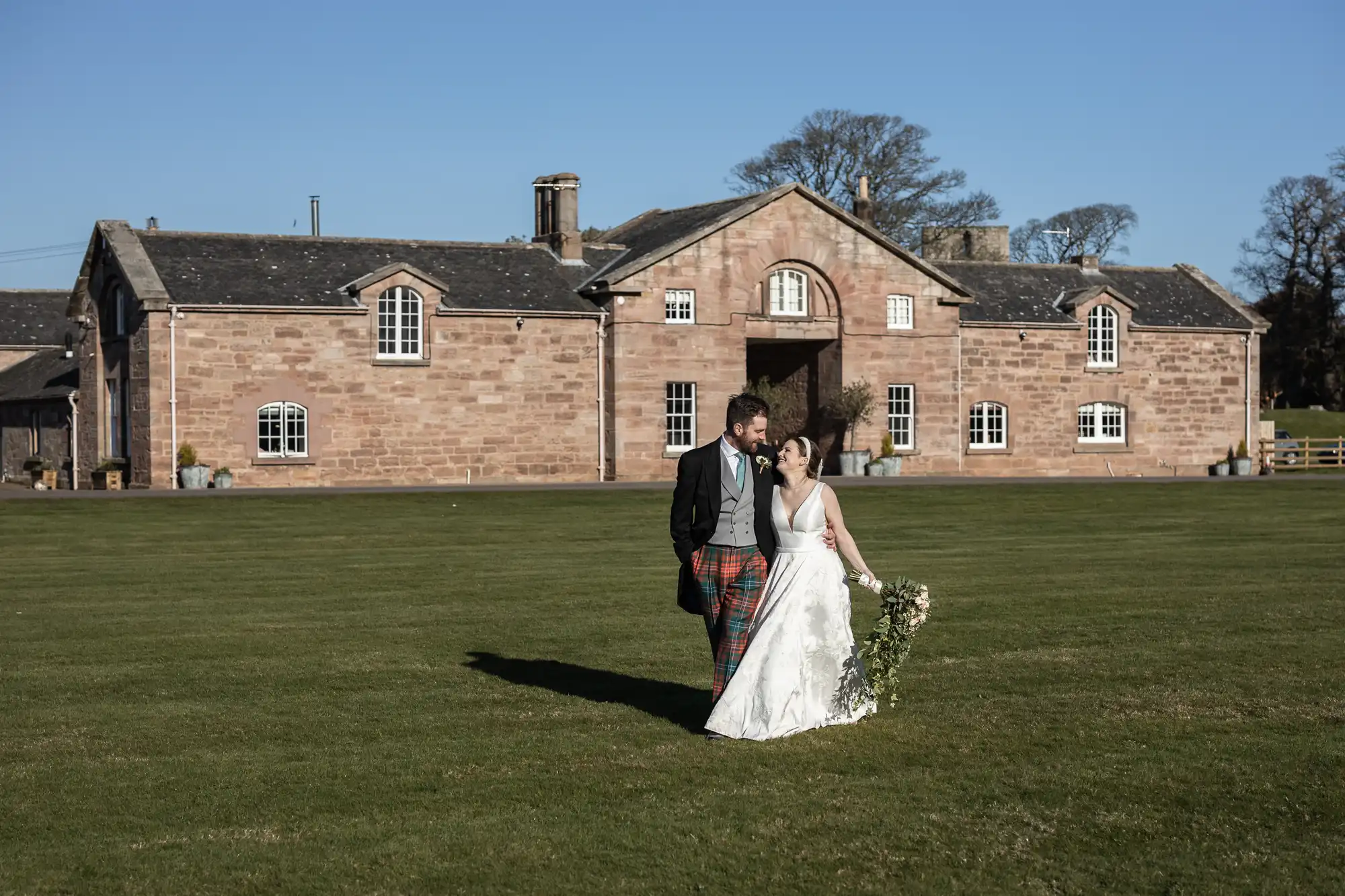 A bride and groom walk hand in hand on a lawn in front of a large, historic brick building on a sunny day. The bride wears a white dress and holds a bouquet, while the groom wears a suit with tartan pants.