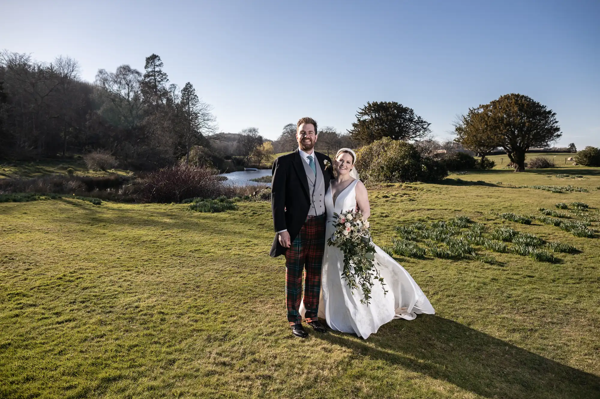 A couple dressed in formal attire stands on a grassy field near a body of water, with trees in the background. The man is wearing a suit with plaid trousers, and the woman is in a white wedding dress holding a bouquet.