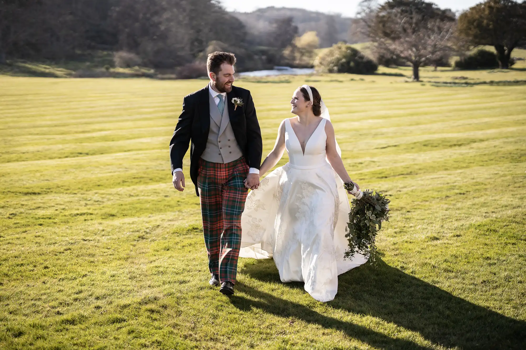 A bride in a white gown and a groom in a suit with plaid pants walk hand in hand on a green lawn, smiling at each other.