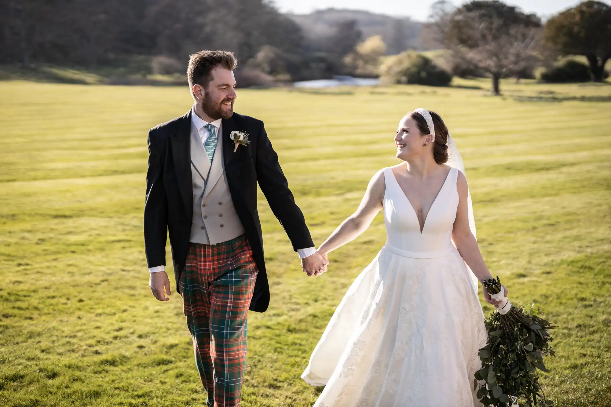 A bride in a white dress and a groom in a tartan kilt and morning coat hold hands and smile at each other while walking on a grassy field.