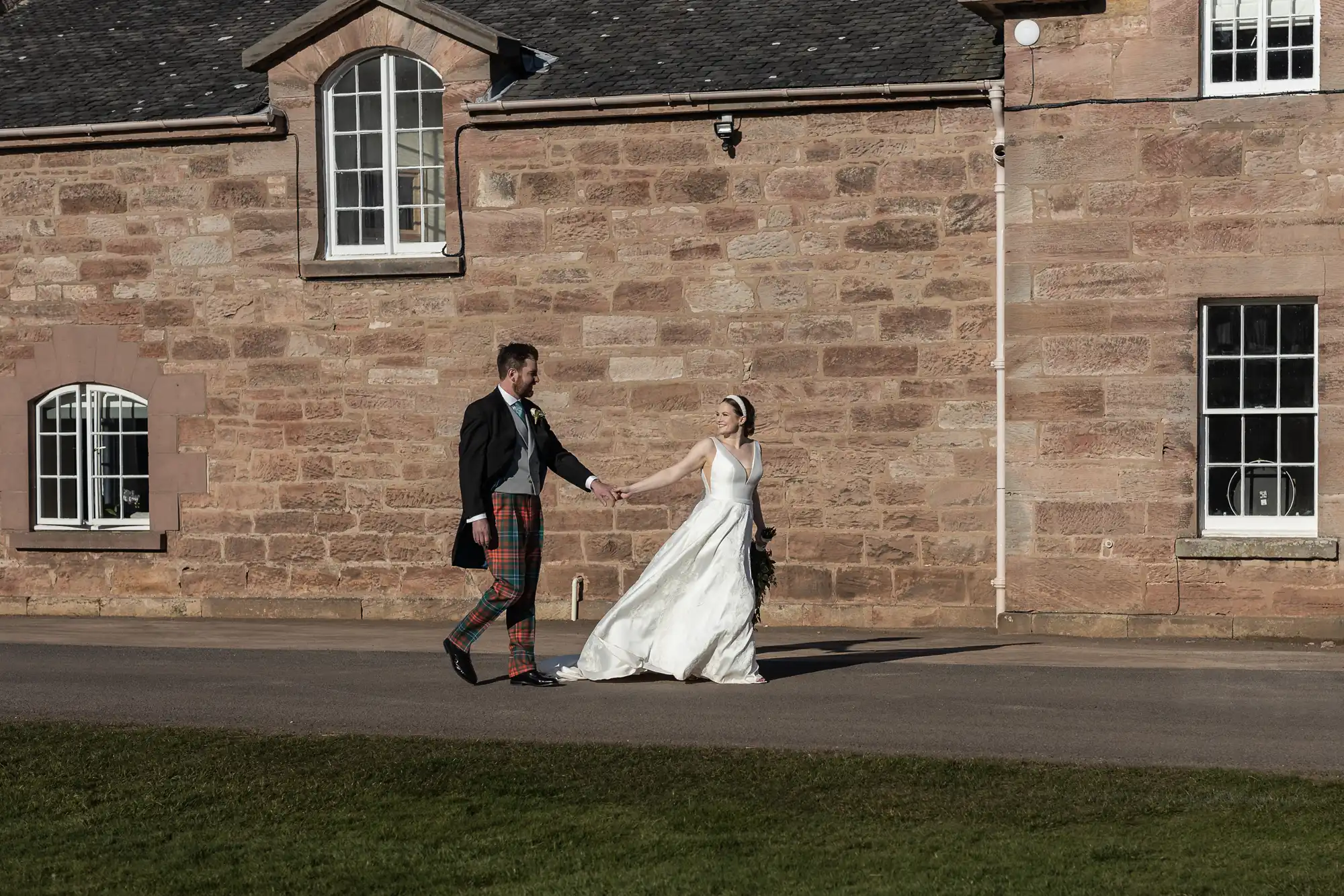 A bride and groom walk hand in hand outside a stone building. The groom wears a suit jacket with tartan trousers, and the bride is in a white dress.