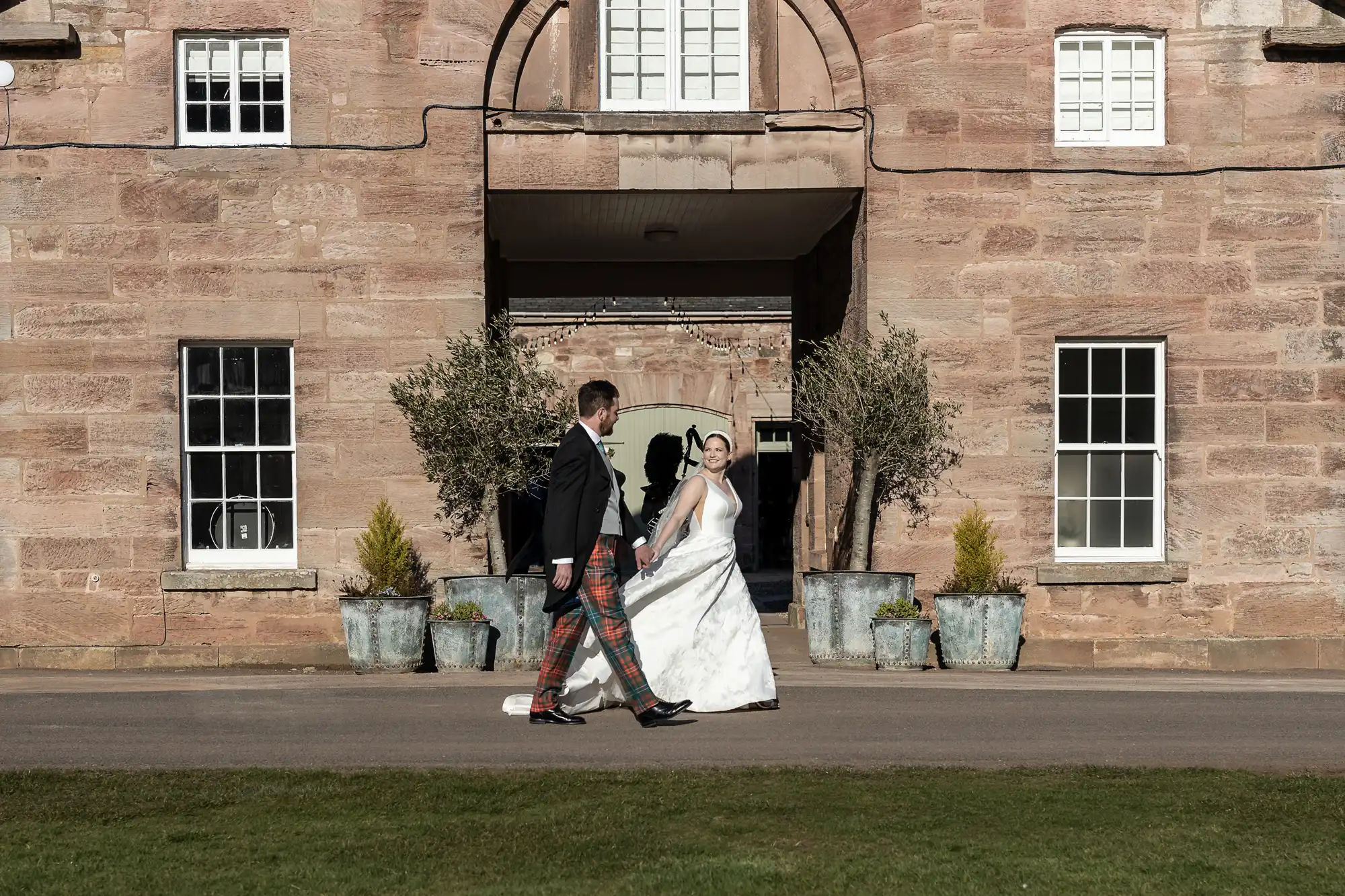 A bride and groom walk in front of a large stone building with white-framed windows and potted plants. The groom is in a suit, and the bride is in a white wedding dress.