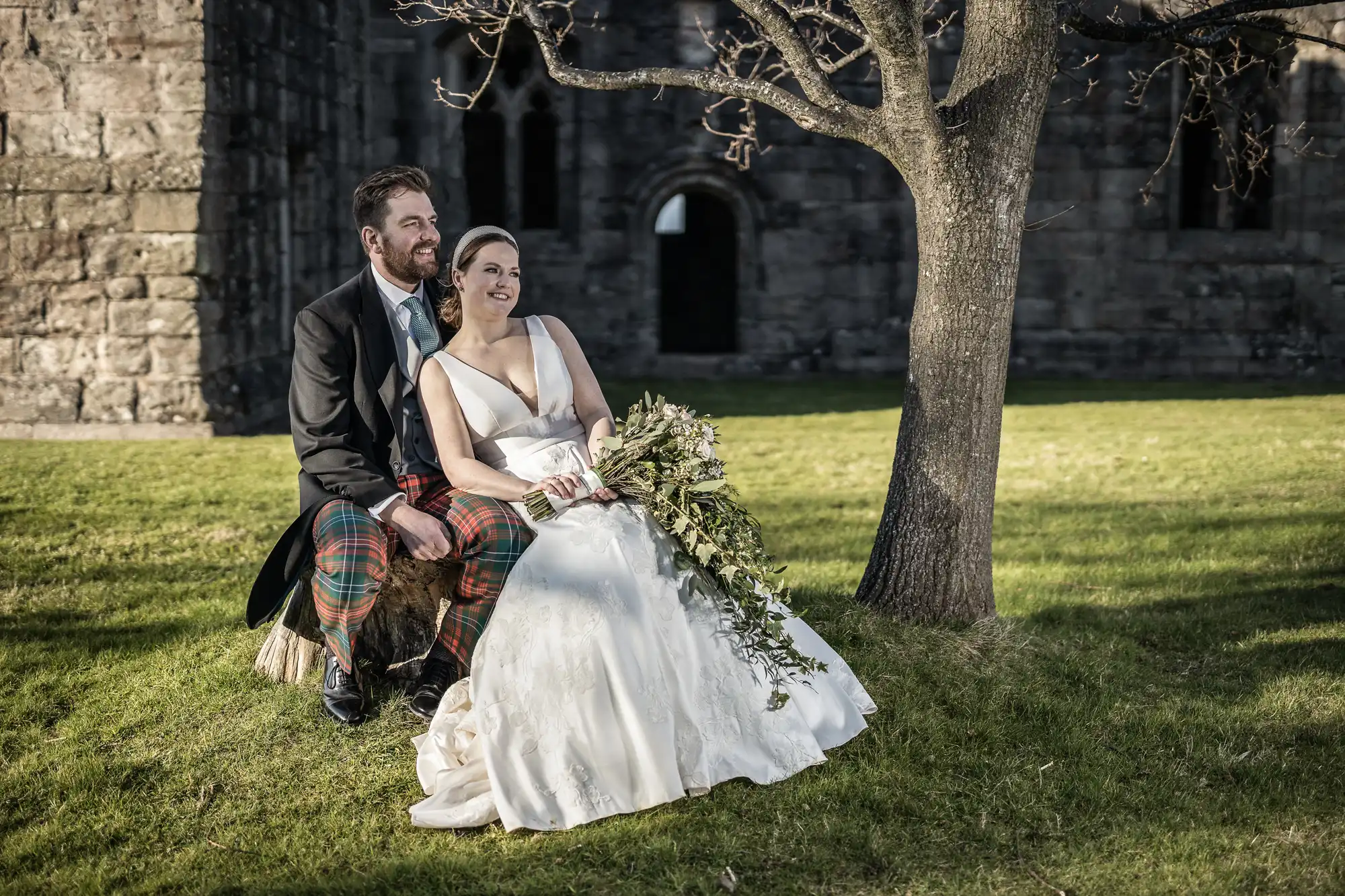 A bride in a white dress and groom in a suit with a kilt sit under a tree, smiling and looking into the distance at an outdoor wedding beside a stone building.