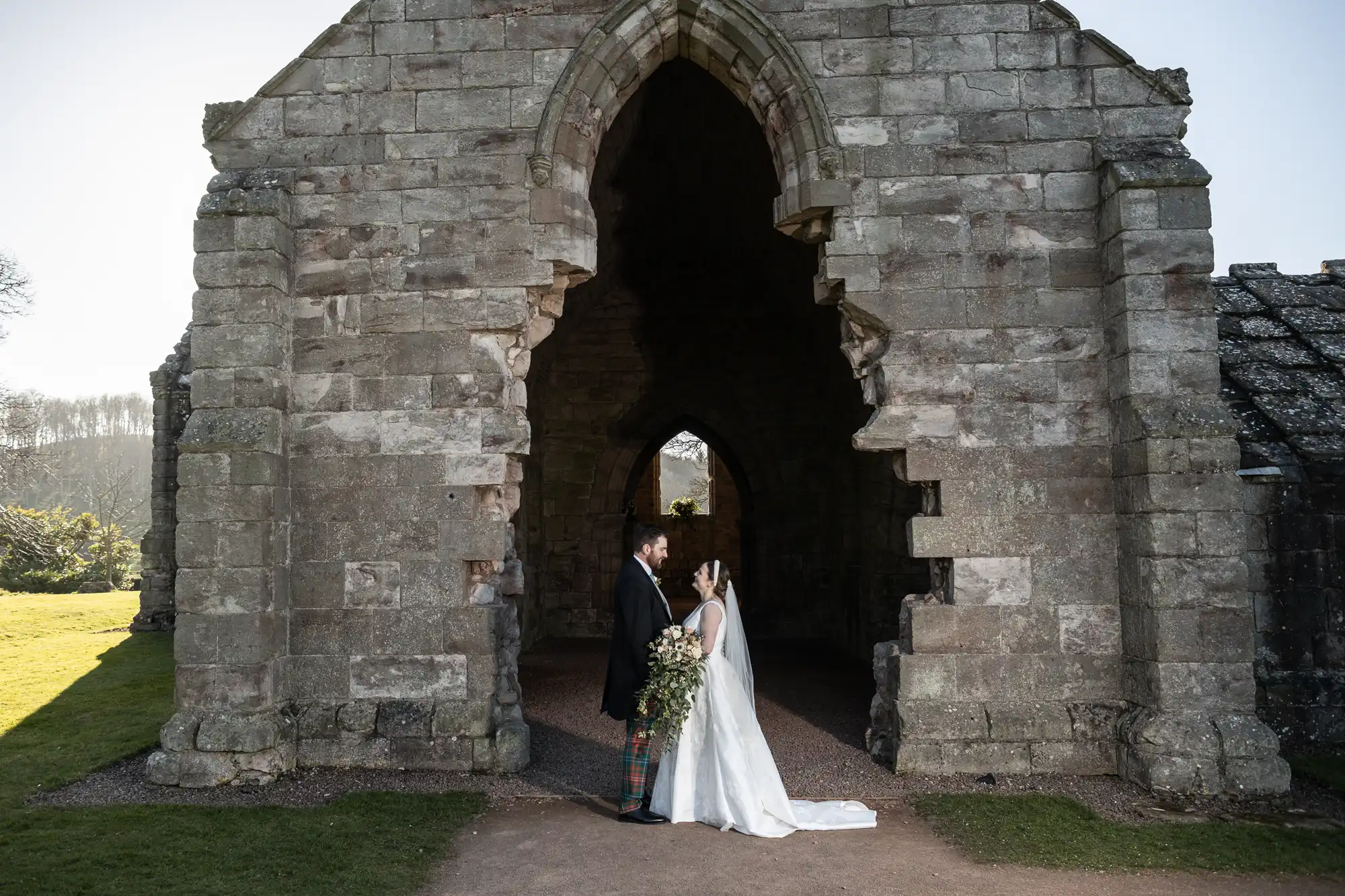 A bride and groom stand facing each other, holding hands in front of an ancient stone archway with a large opening and rugged stone walls. The bride wears a white dress and the groom a dark suit with a tartan sash.