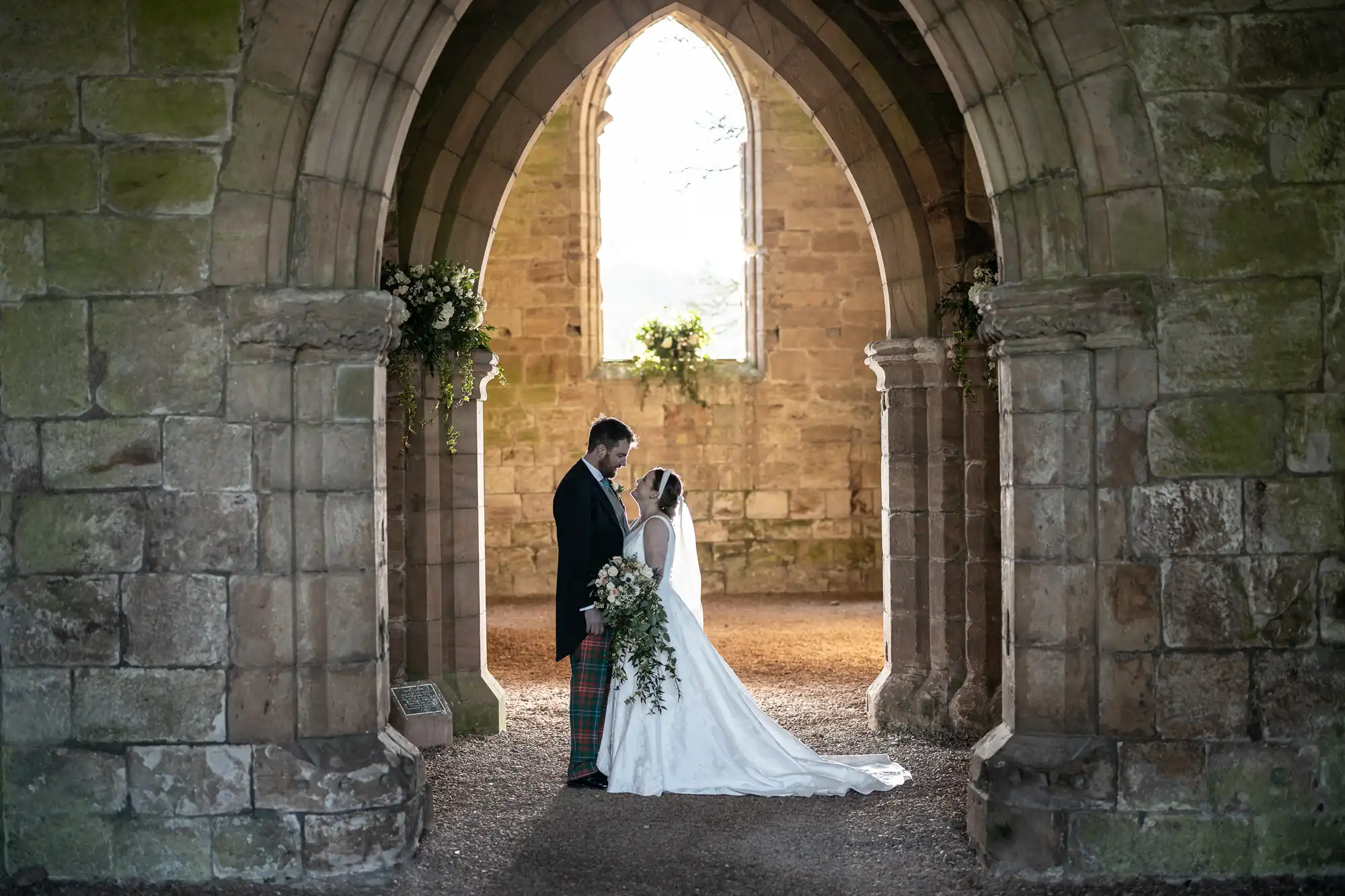 A couple dressed in wedding attire stand under a stone archway, facing each other. The bride holds a bouquet of flowers, and they are framed by the architectural structure of an old building.