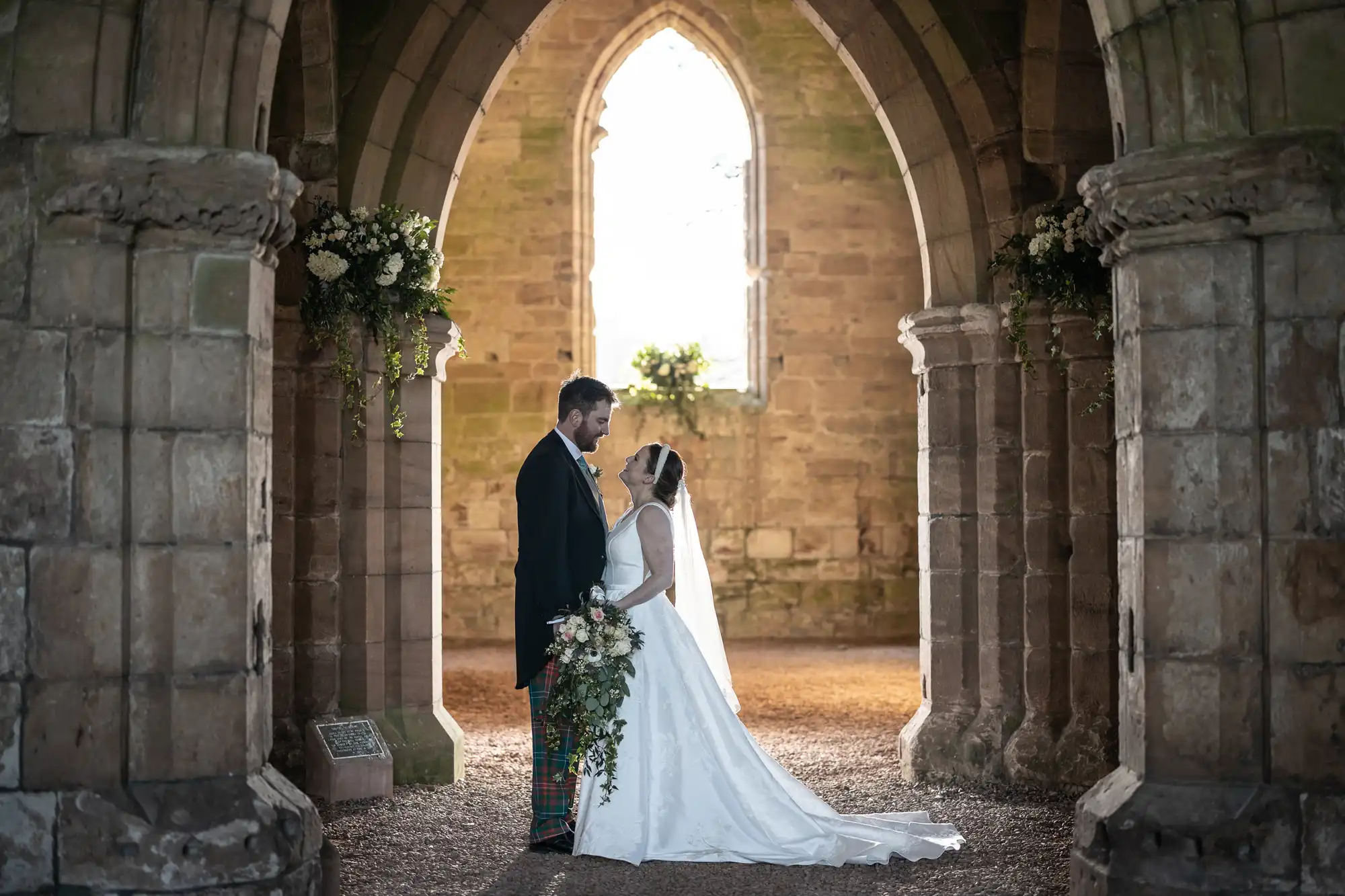 A bride and groom stand together in an ancient stone archway, gazing into each other's eyes. The bride holds a cascading bouquet of flowers. Sunlight filters through a window behind them.