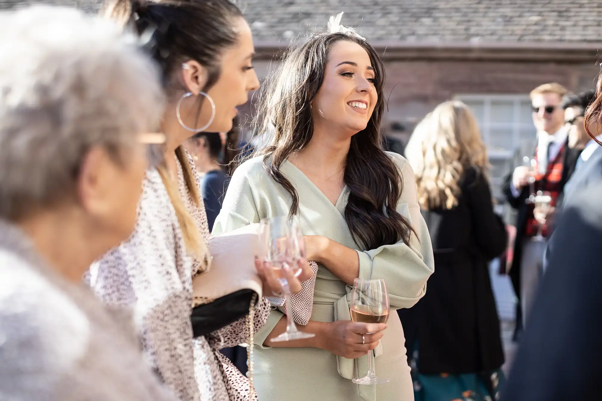 A group of people, including a young woman in a light-colored dress holding a glass, are engaged in conversation at a social event. The background shows more attendees and a brick building.