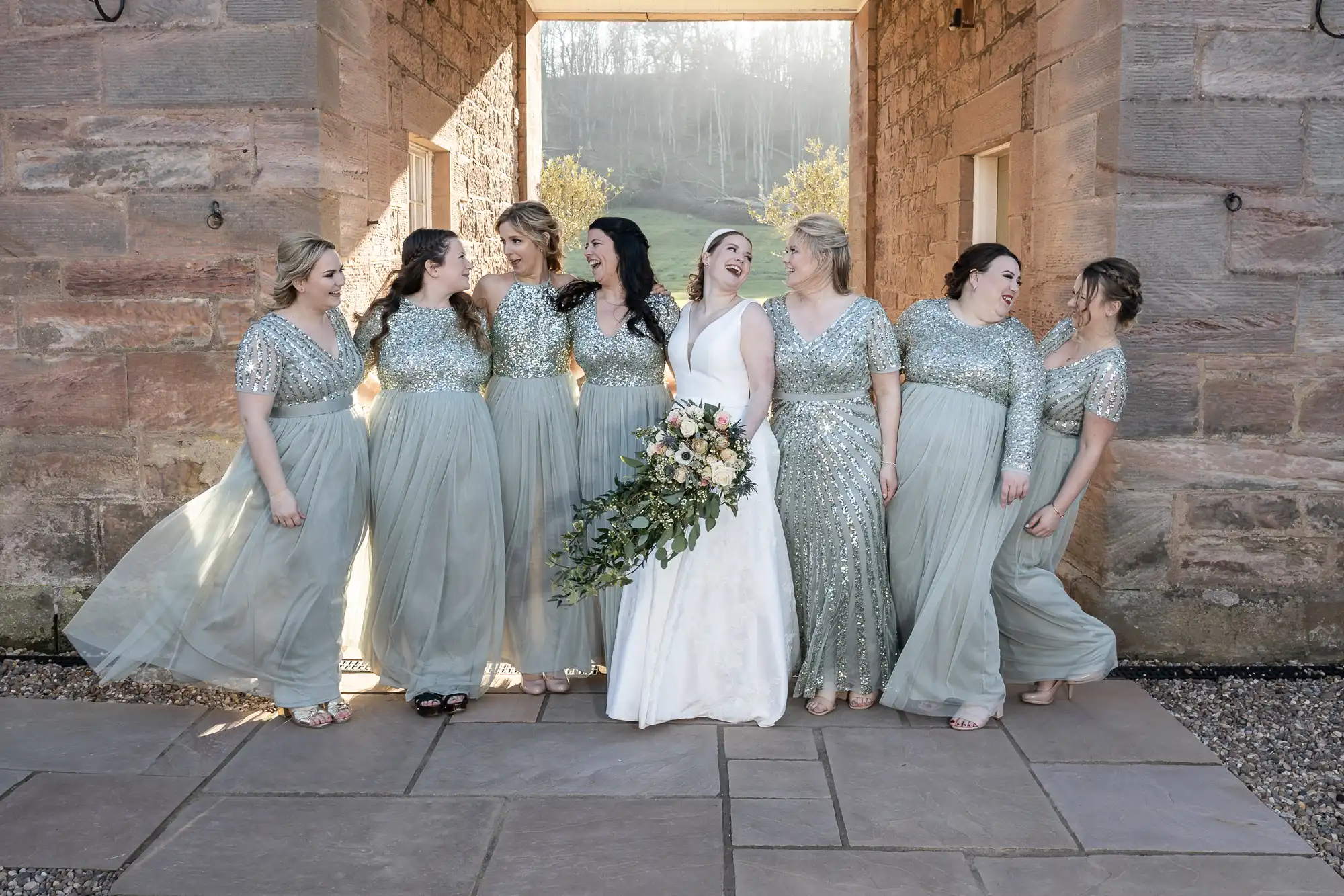 A bride in a white dress stands with seven bridesmaids in light green dresses. They are posed in a bright, sunlit stone corridor. The bride holds a large bouquet of flowers.