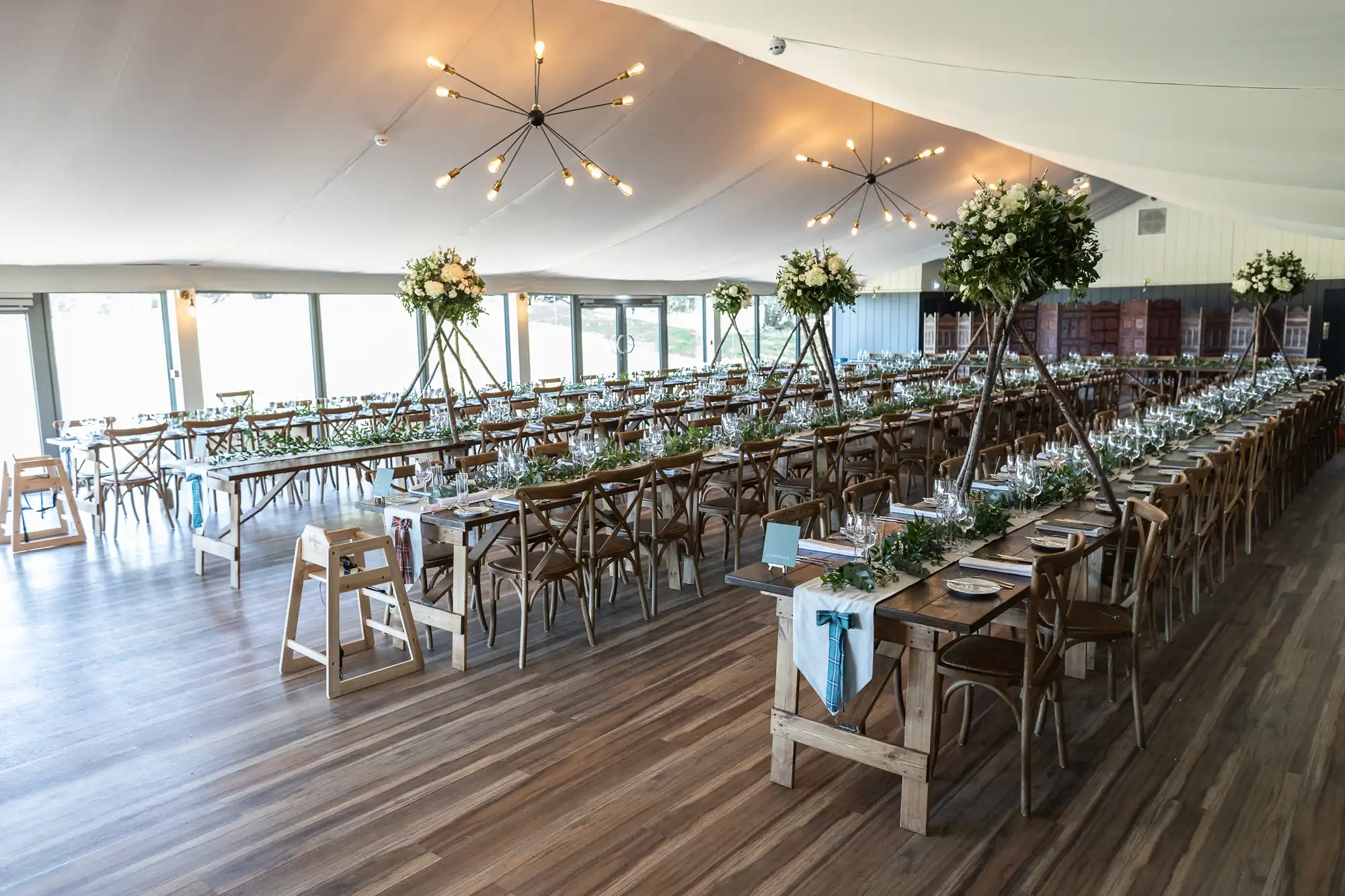 A large banquet hall set up for an event with long wooden tables and chairs, decorated with tall floral centerpieces and table settings. The space features large windows and contemporary lighting.