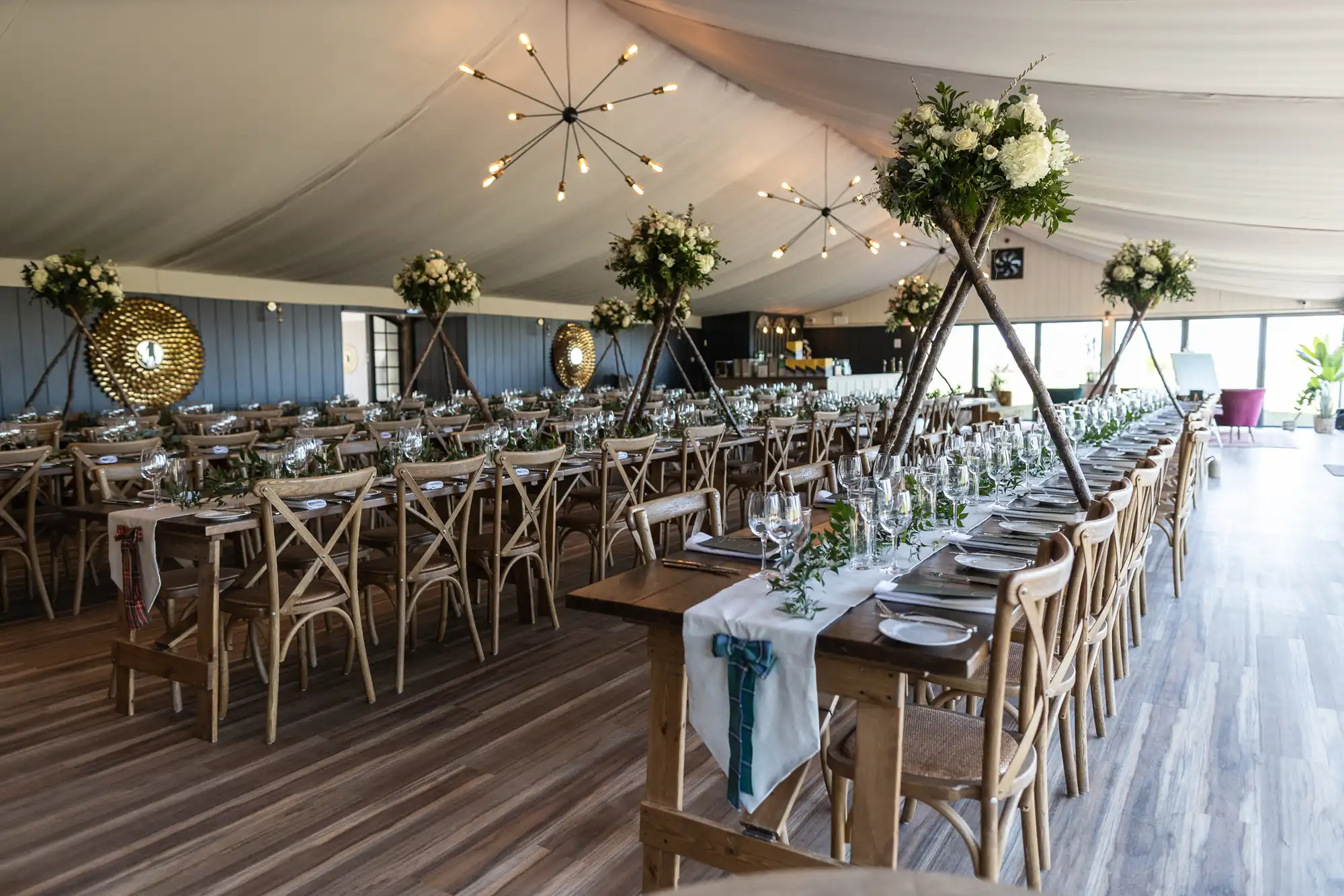 A banquet hall with long wooden tables set for a formal event. Each table is adorned with green foliage, white flowers, and elegant glassware. Suspended light fixtures hang from the ceiling.