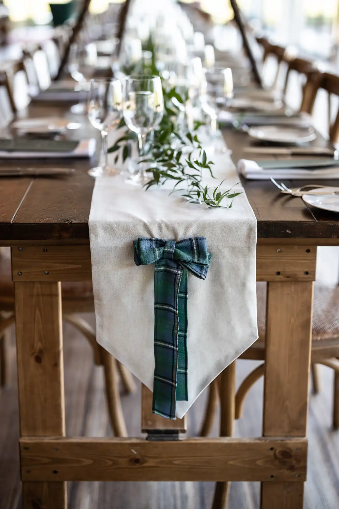 A long, wooden dining table is adorned with a white table runner and decorations, including a blue-green plaid bow and green foliage, set with wine glasses, plates, and cutlery.