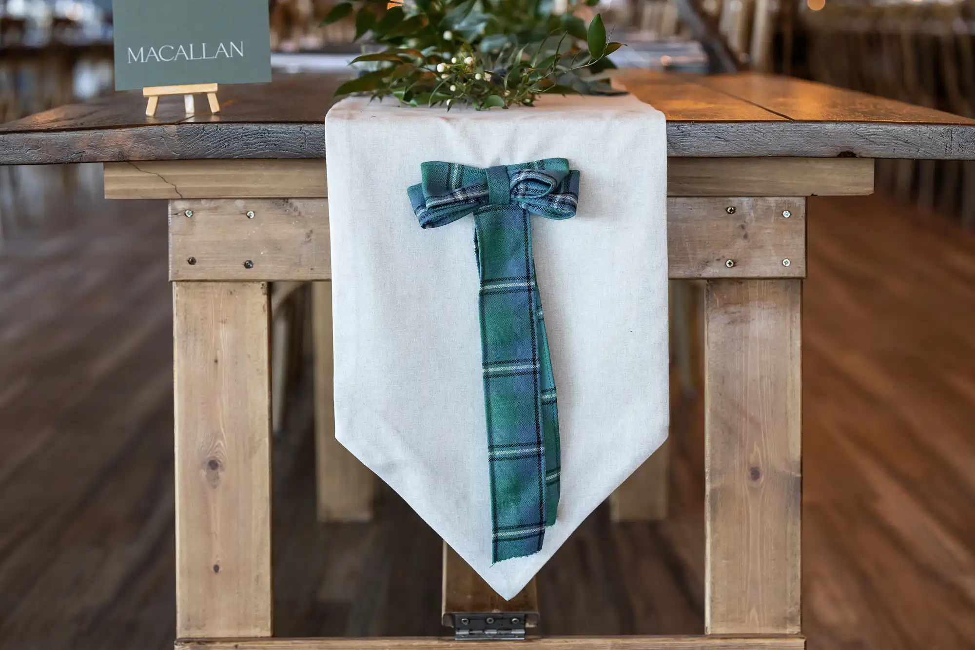 A wooden table decorated with a white runner and a green plaid bow. A small sign with "Macallan" written on it is on the left corner of the table.