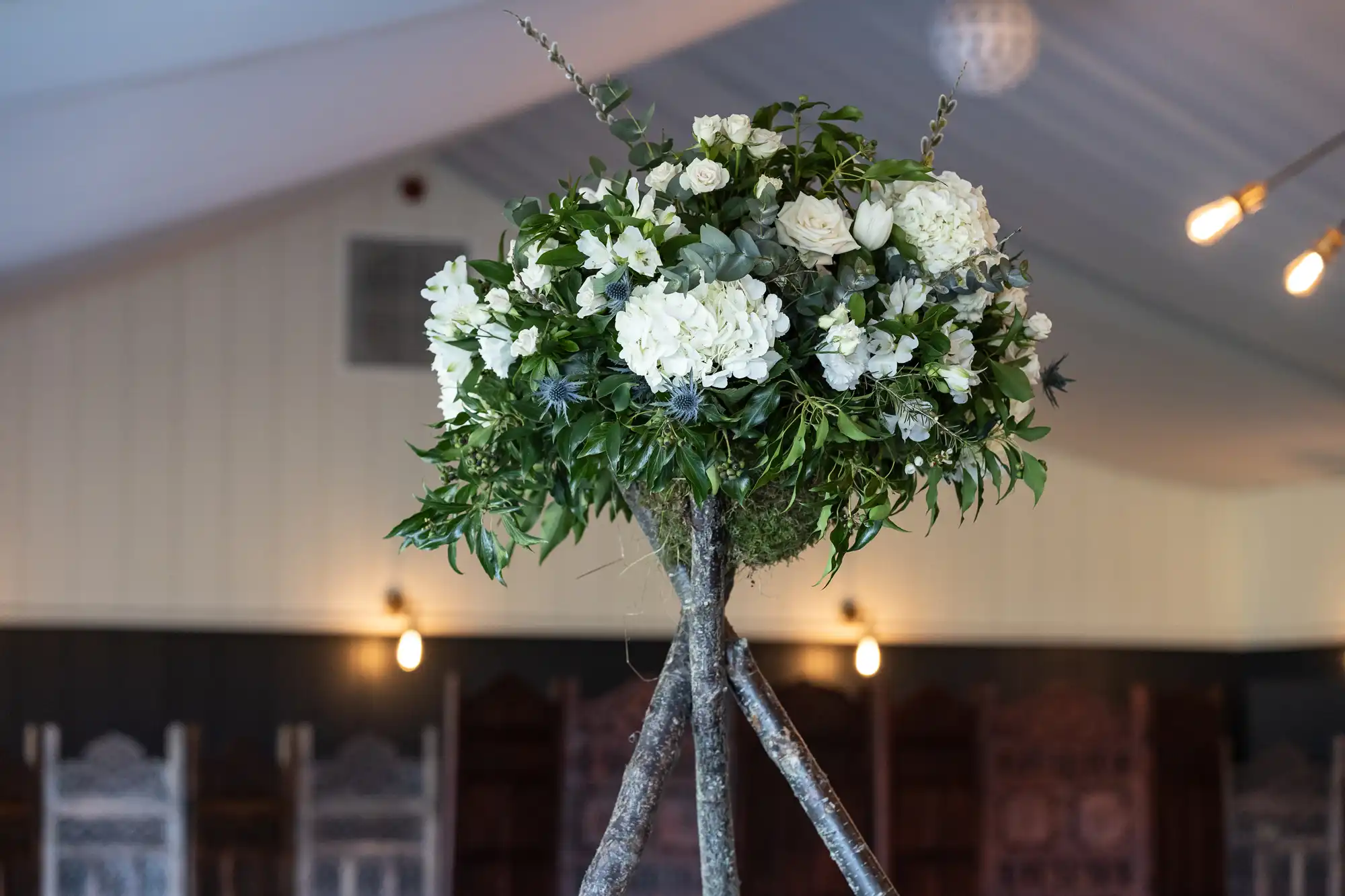 A tall arrangement of white flowers and greenery on a rustic wooden tripod stand, placed in a softly lit room with high ceilings.