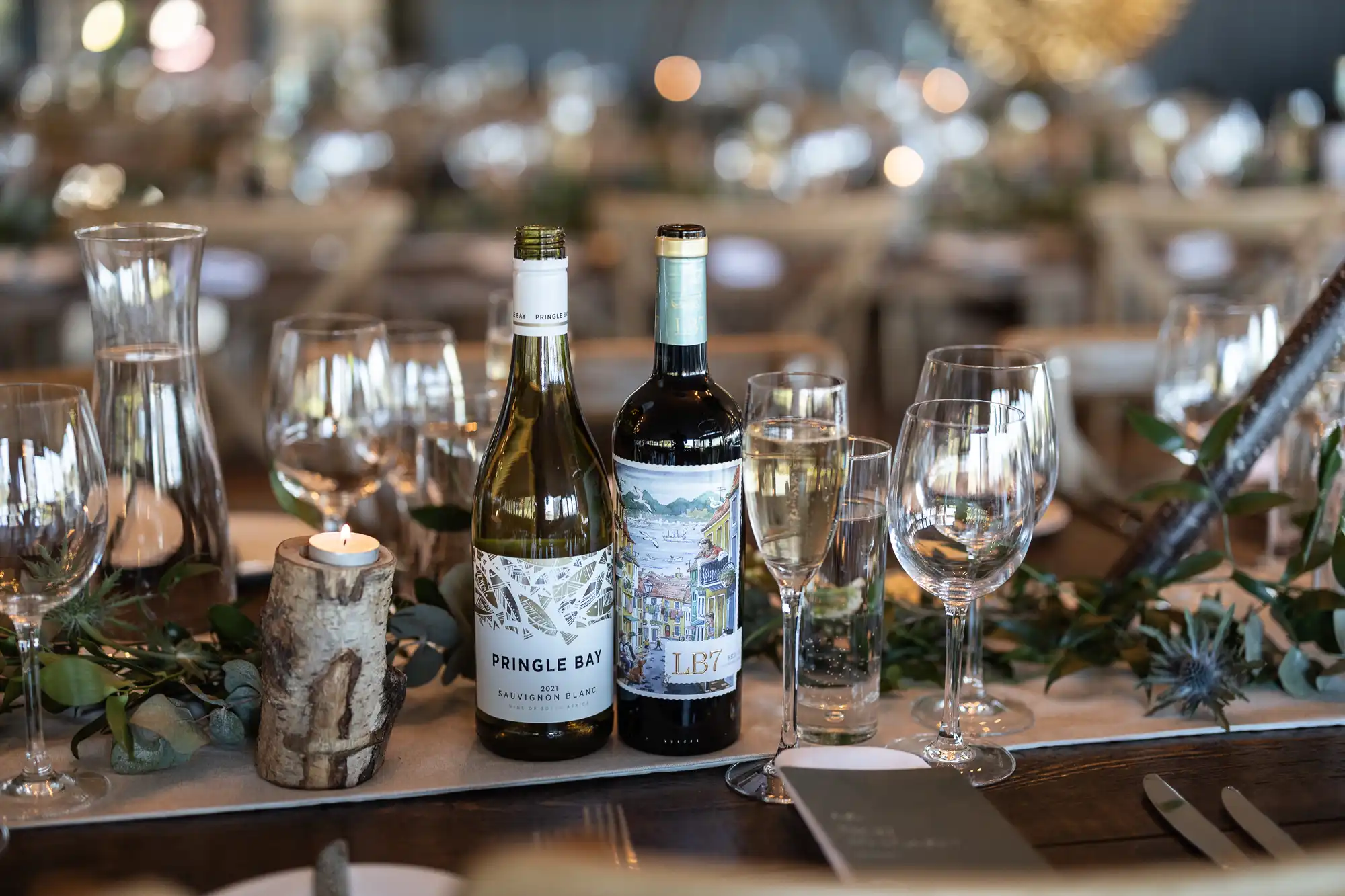 A table setting featuring two bottles of wine, numerous wine glasses, a lit candle in a rustic holder, and greenery garland decor. The background shows more tables set similarly.