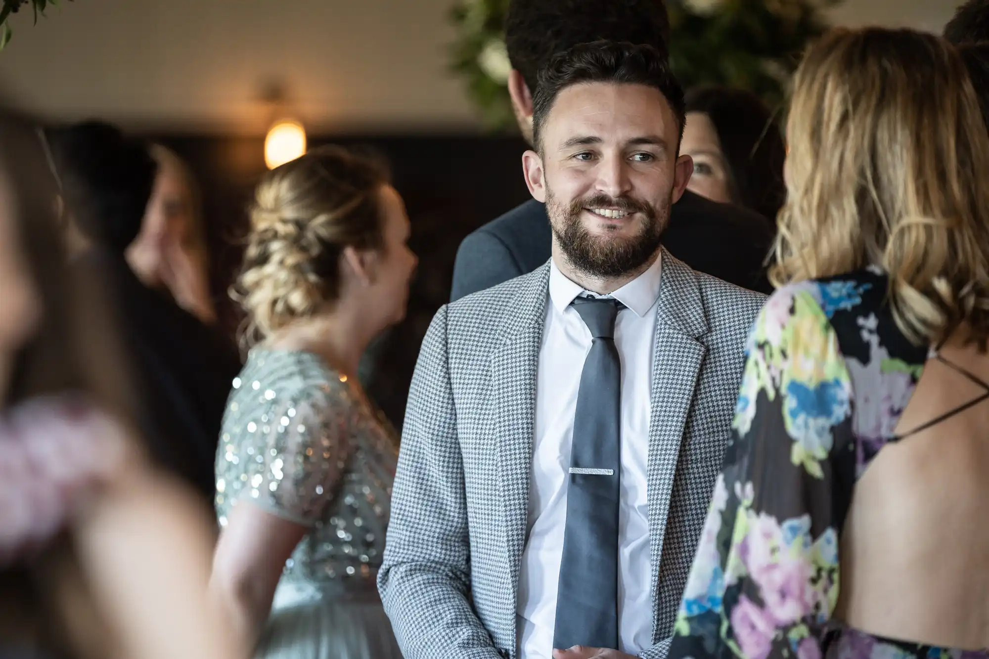 A bearded man in a gray suit and tie stands indoors, smiling and engaging with a woman in a floral dress among a crowd of people.