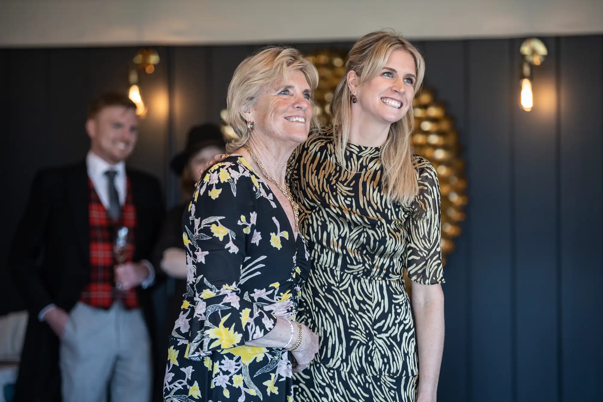 Two women, one older and one younger, stand closely together, smiling. The older woman wears a floral dress, and the younger woman wears a zebra-print dress. Blurred people are in the background.