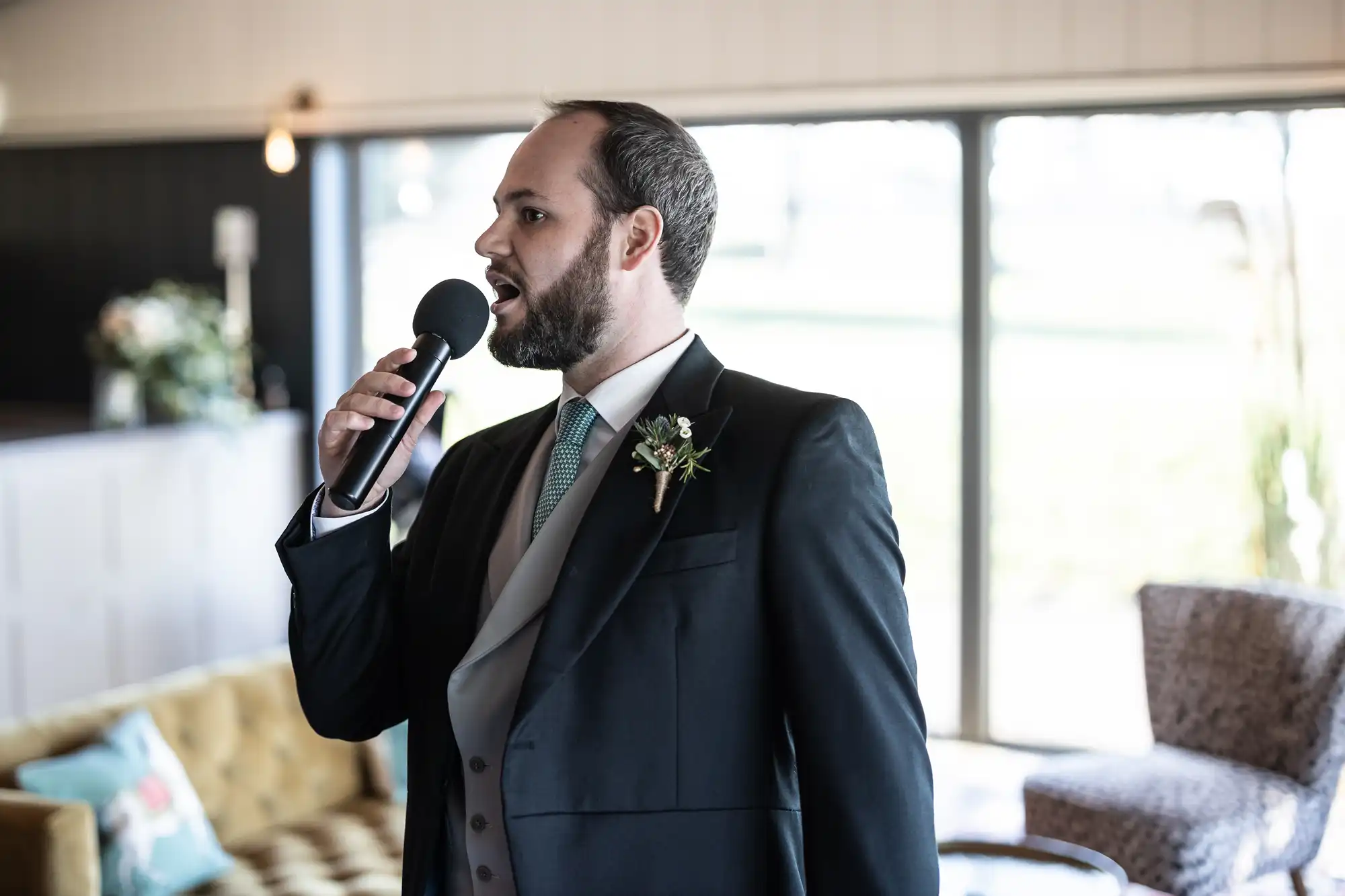 A man in a suit speaks into a microphone, standing indoors near a window with bright natural light.