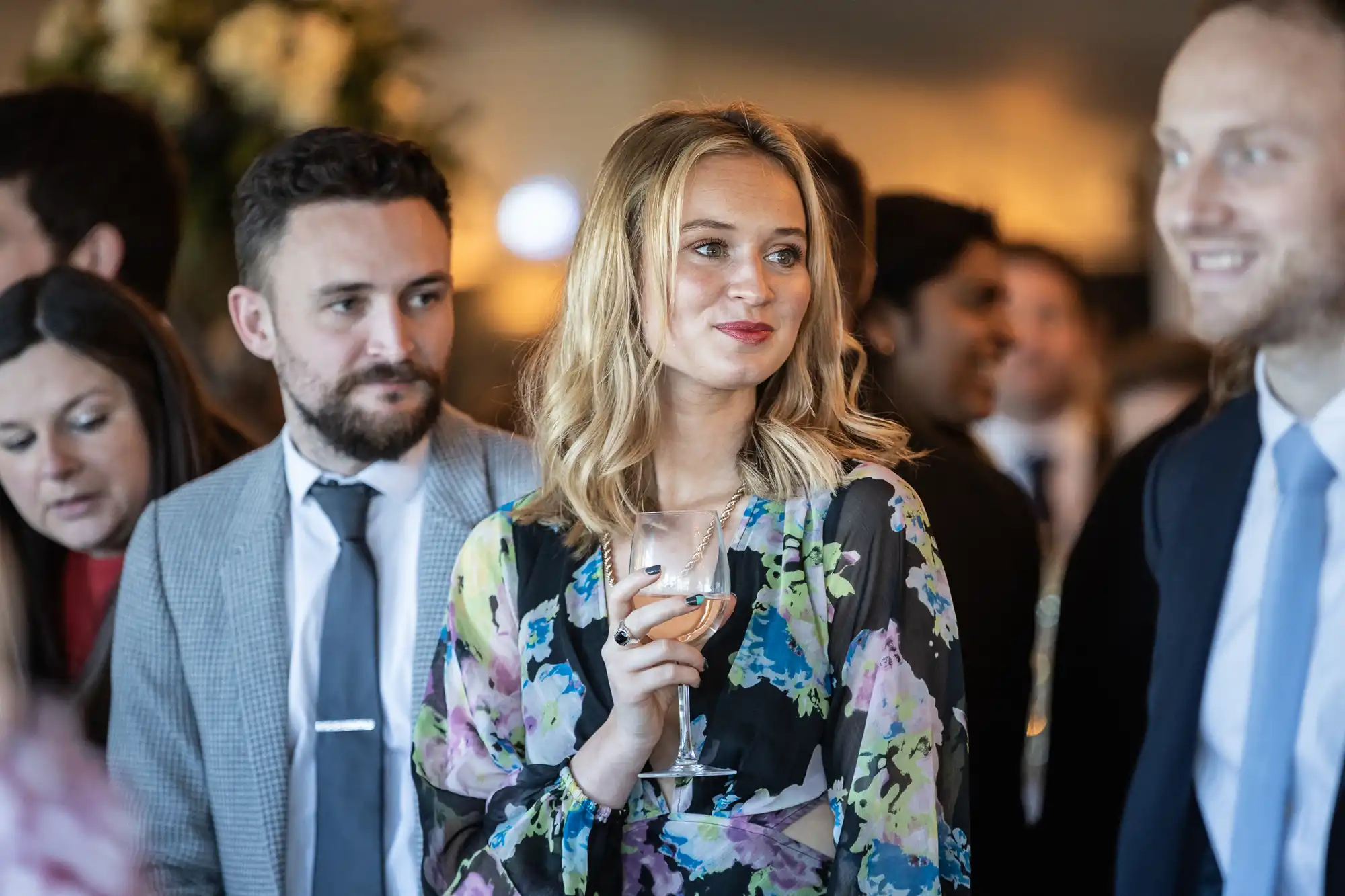 A woman holding a wine glass stands among a group of people dressed in formal attire at a social event.