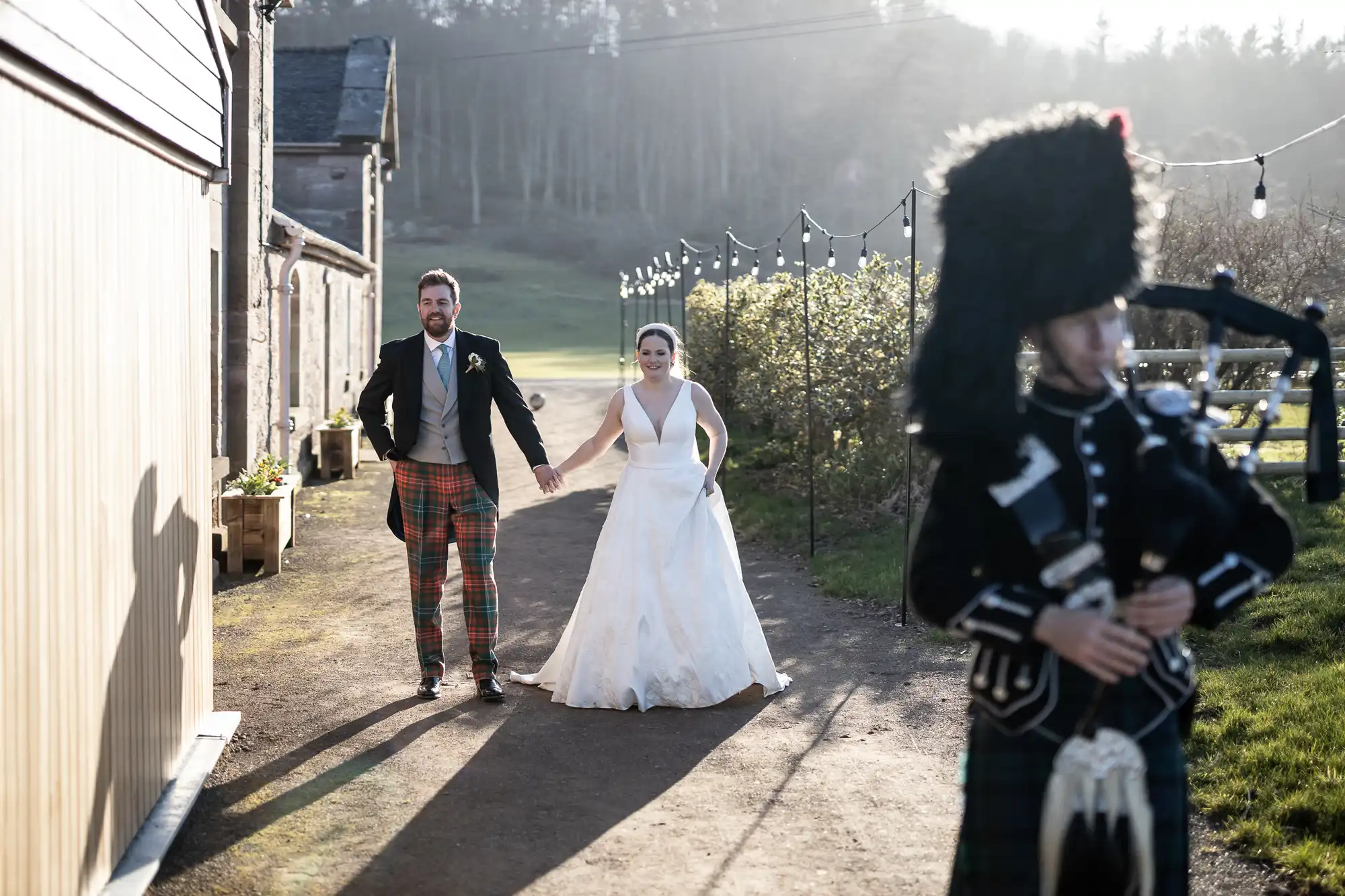 Bride and groom walking outside holding hands, groom in tartan pants and bride in white gown, bagpiper in traditional attire plays in the foreground.