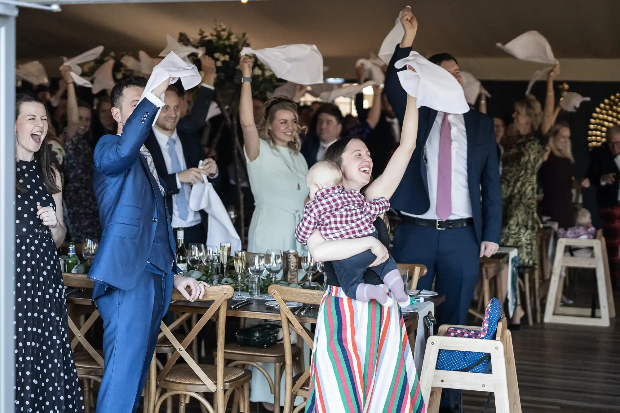 A group of people, some standing and some seated, enthusiastically waving white napkins in the air. A woman holds a baby in the foreground. The setting appears to be a celebratory event or gathering.