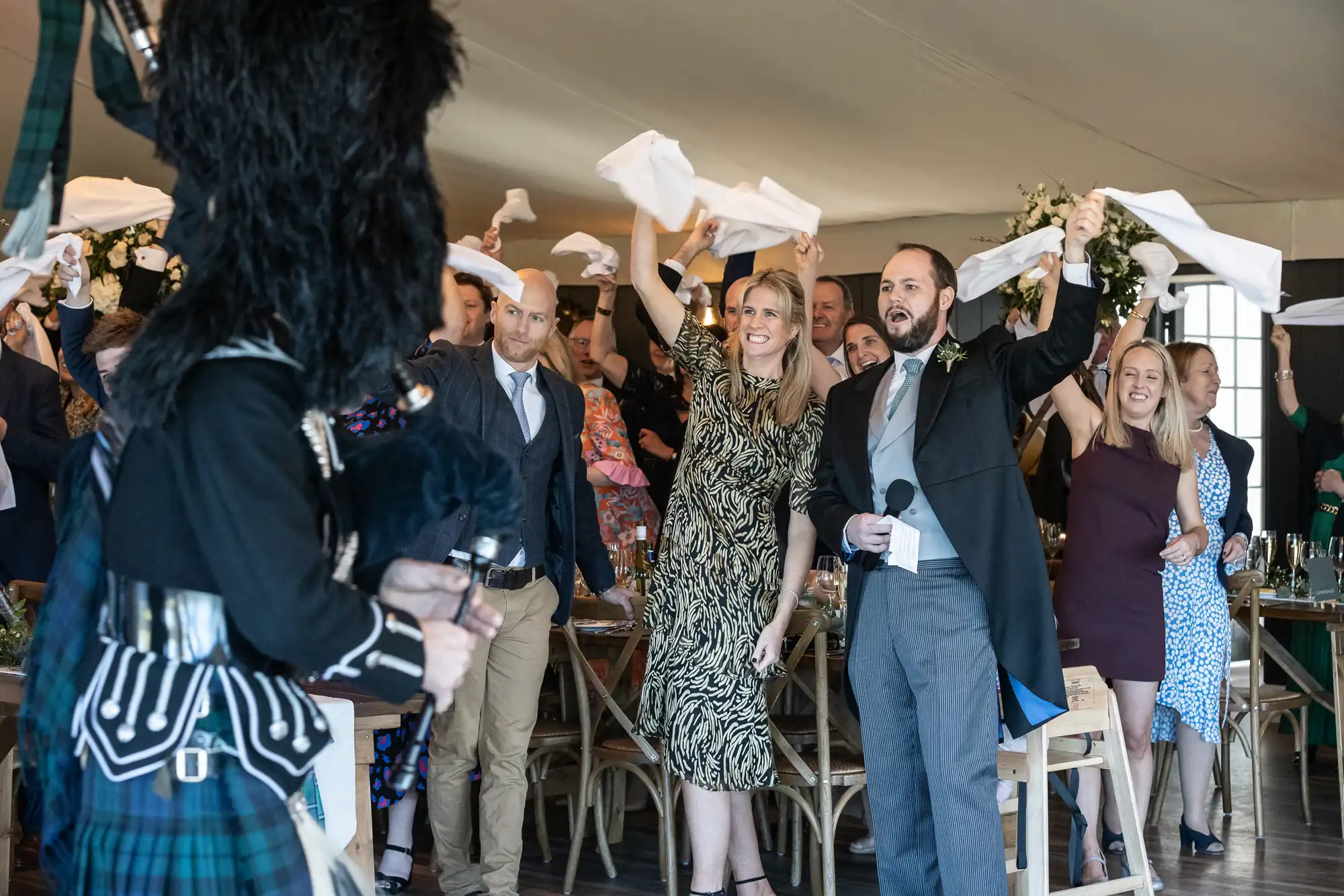 A group of people at a celebration wave napkins while a person in a plaid outfit and tall hat, possibly a performer, stands in the foreground.