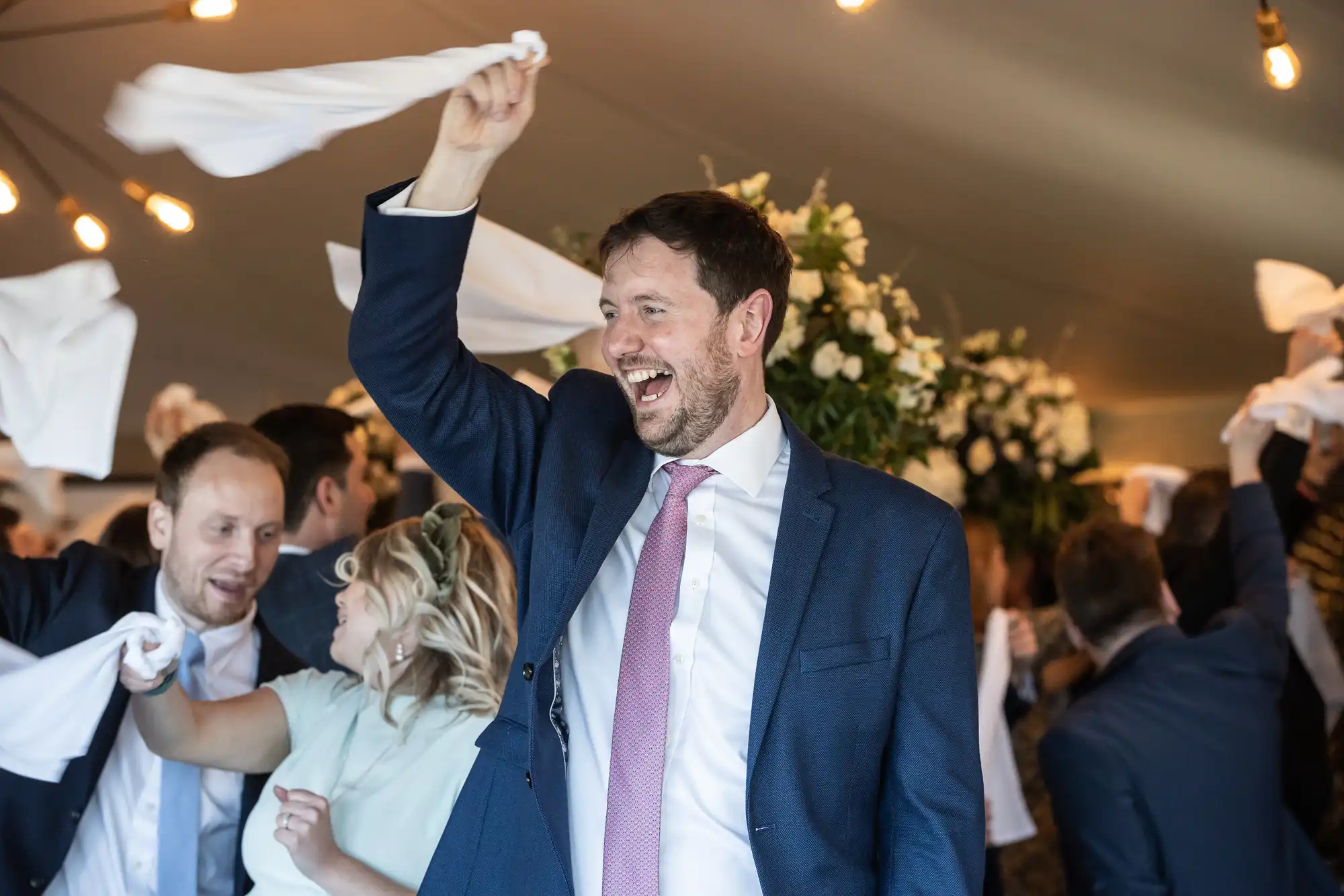 People are joyfully waving white cloths indoors, with one man in the foreground wearing a navy suit and pink tie, smiling and raising his arm.