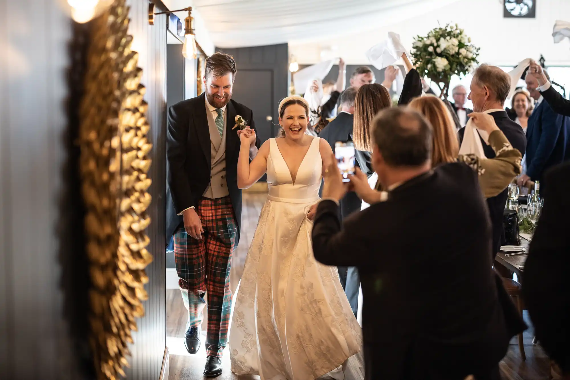 A newlywed couple enters a reception hall, with guests cheering and taking photos. The bride wears a white gown, and the groom wears a suit with tartan trousers.