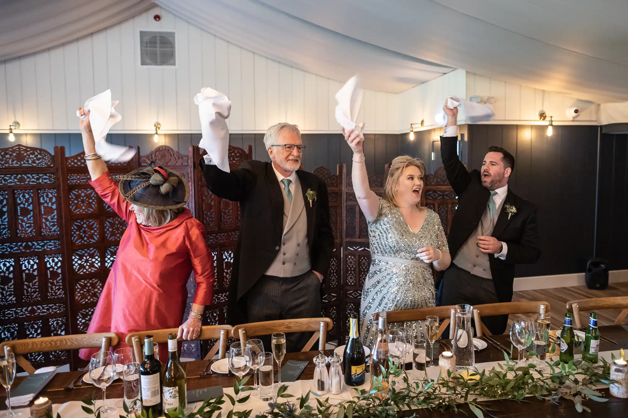 Four people stand behind a table, waving white napkins in the air and smiling, with drinks and table settings visible.