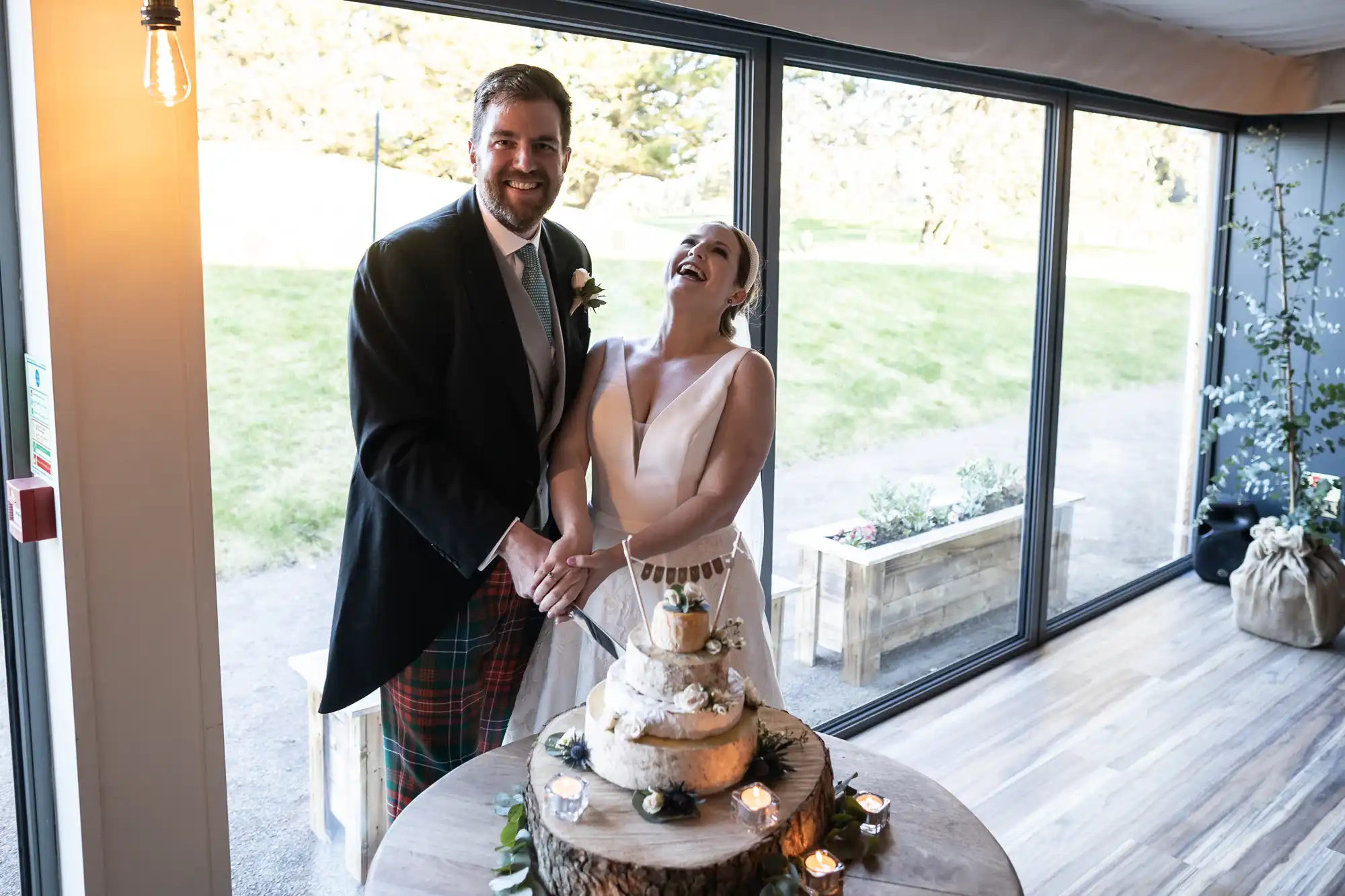 A newlywed couple stands together and smiles while cutting their wedding cake in a bright, modern venue with large windows.