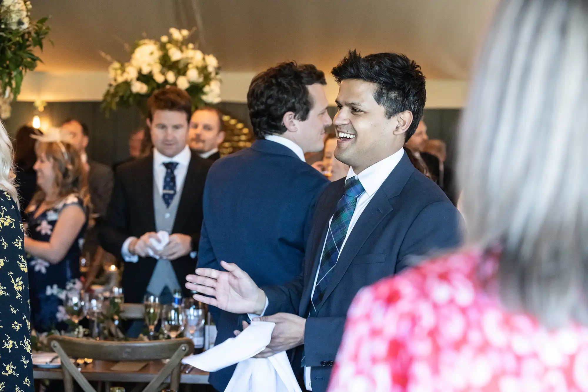 A man in a suit and tie smiling and holding a napkin in a social gathering with people conversing and floral decorations in the background.