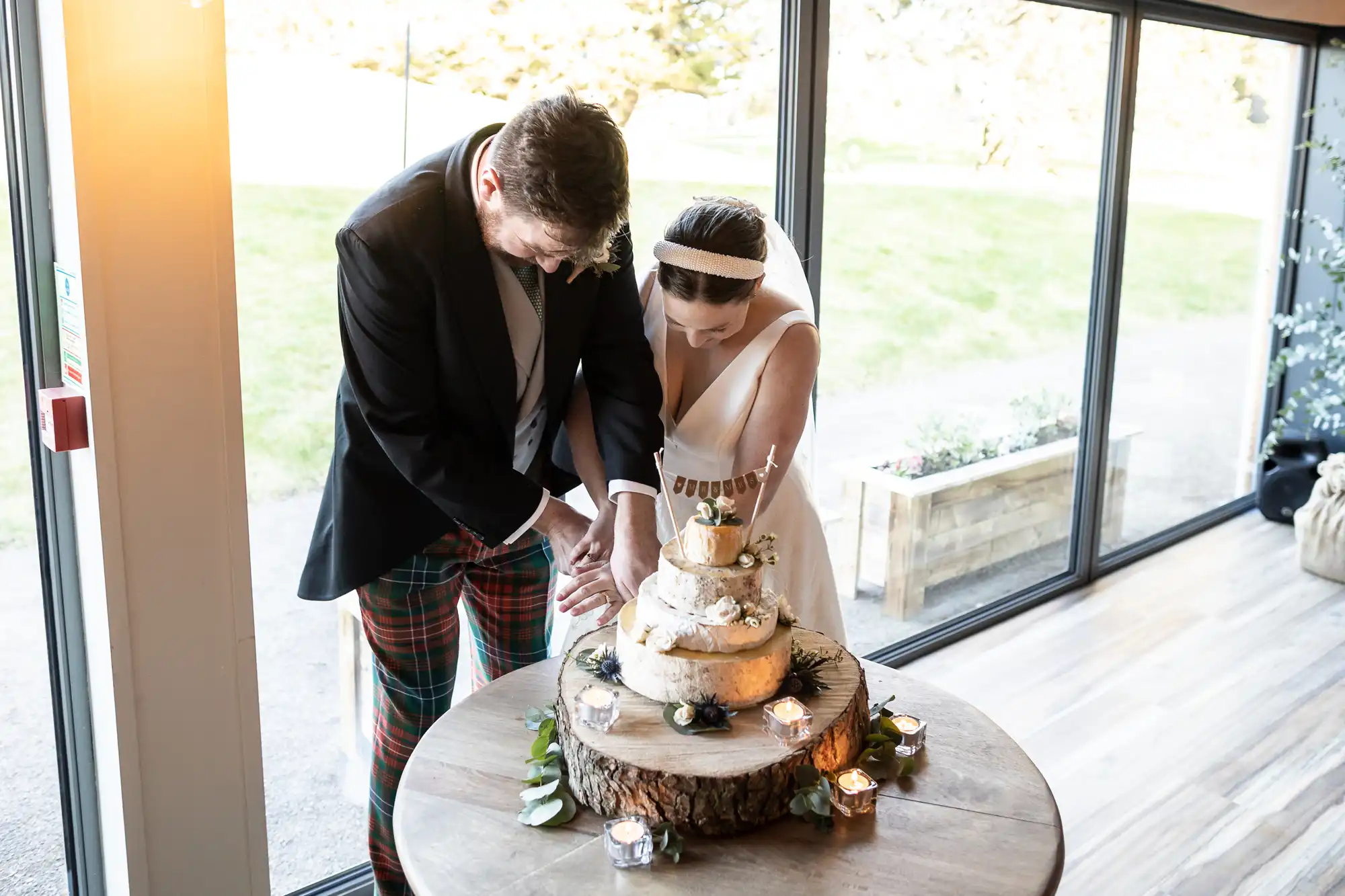 A couple in formal attire jointly cuts a cake on a small wooden table decorated with flowers and candles, set near large windows overlooking a garden.