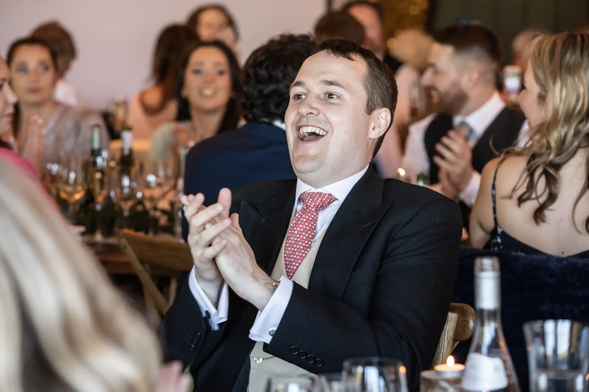 A man in a suit claps and smiles at a formal event, surrounded by people seated at tables with glasses and bottles.