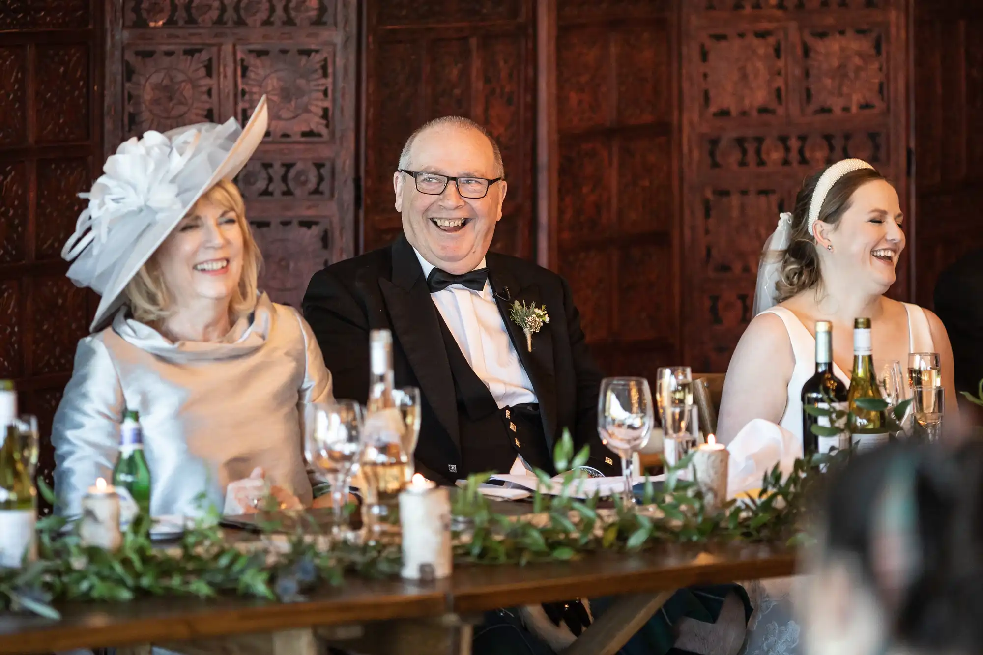 Three people dressed formally are seated at a banquet table, smiling and laughing. The table is decorated with greenery, candles, and various drinks.