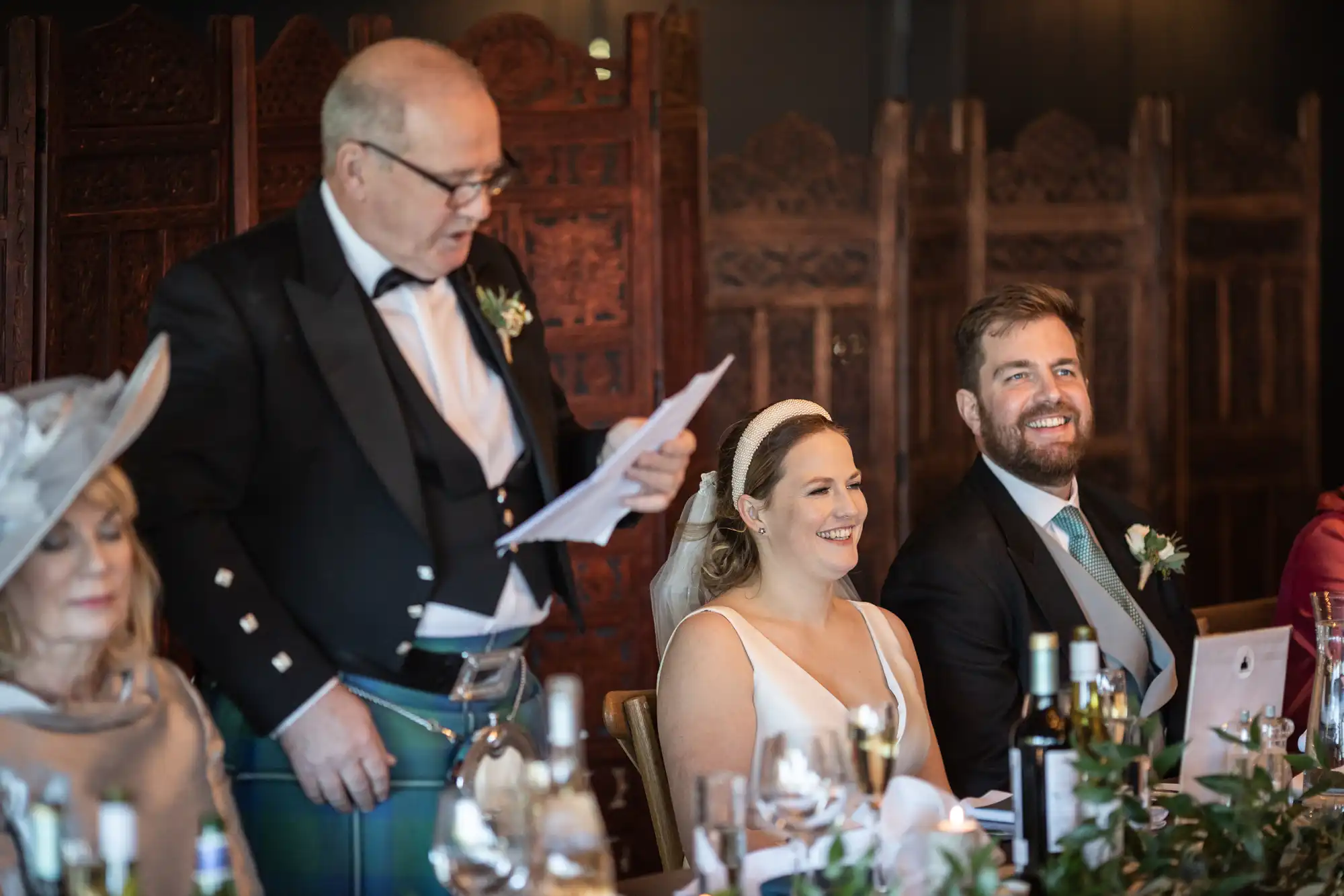 A man in a kilt gives a speech at a wedding reception. The bride, groom, and other guests are seated at a table, smiling and listening attentively.