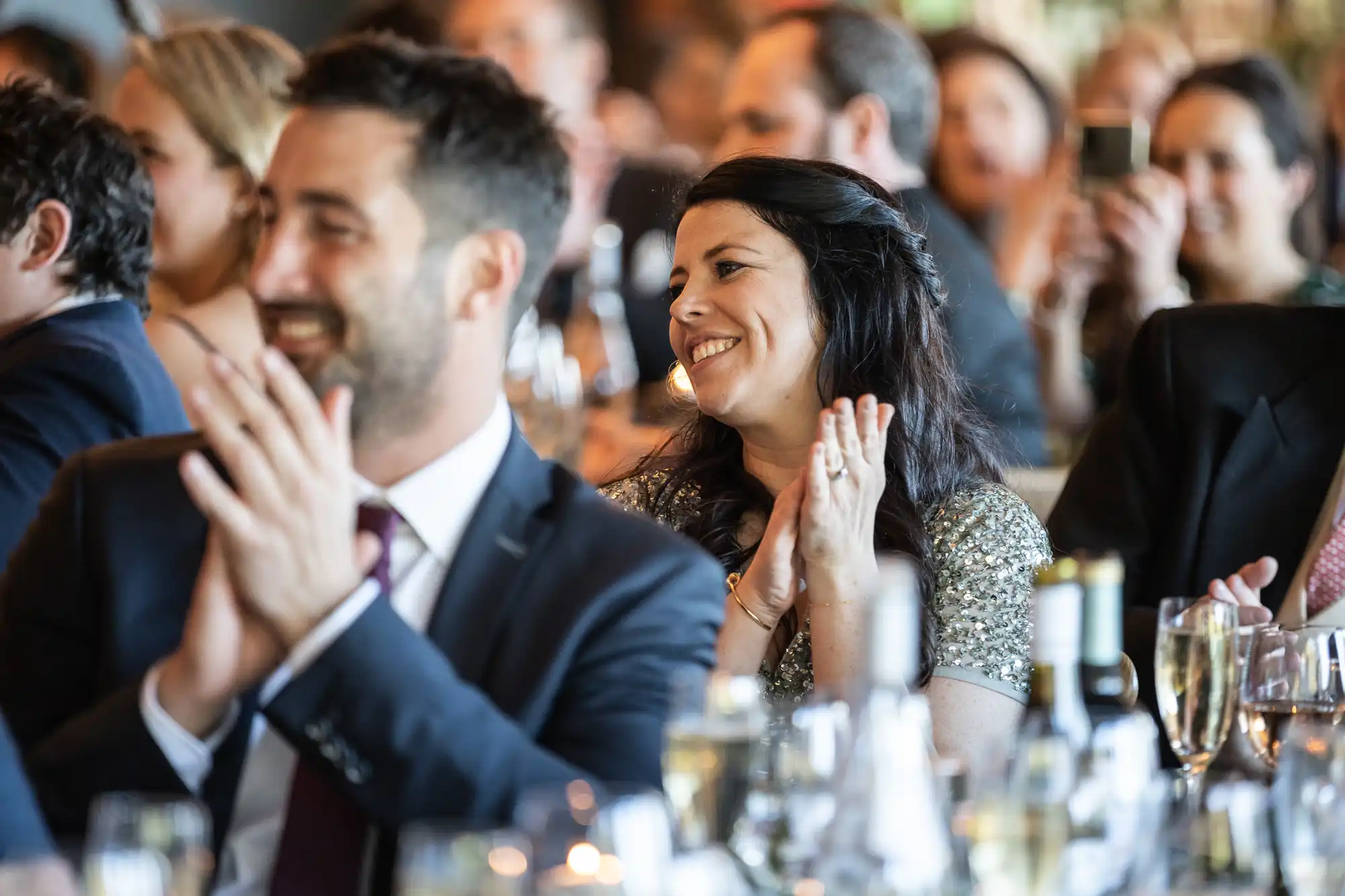 A group of people in formal attire sitting at tables, clapping and smiling, at what appears to be an indoor event or celebration.