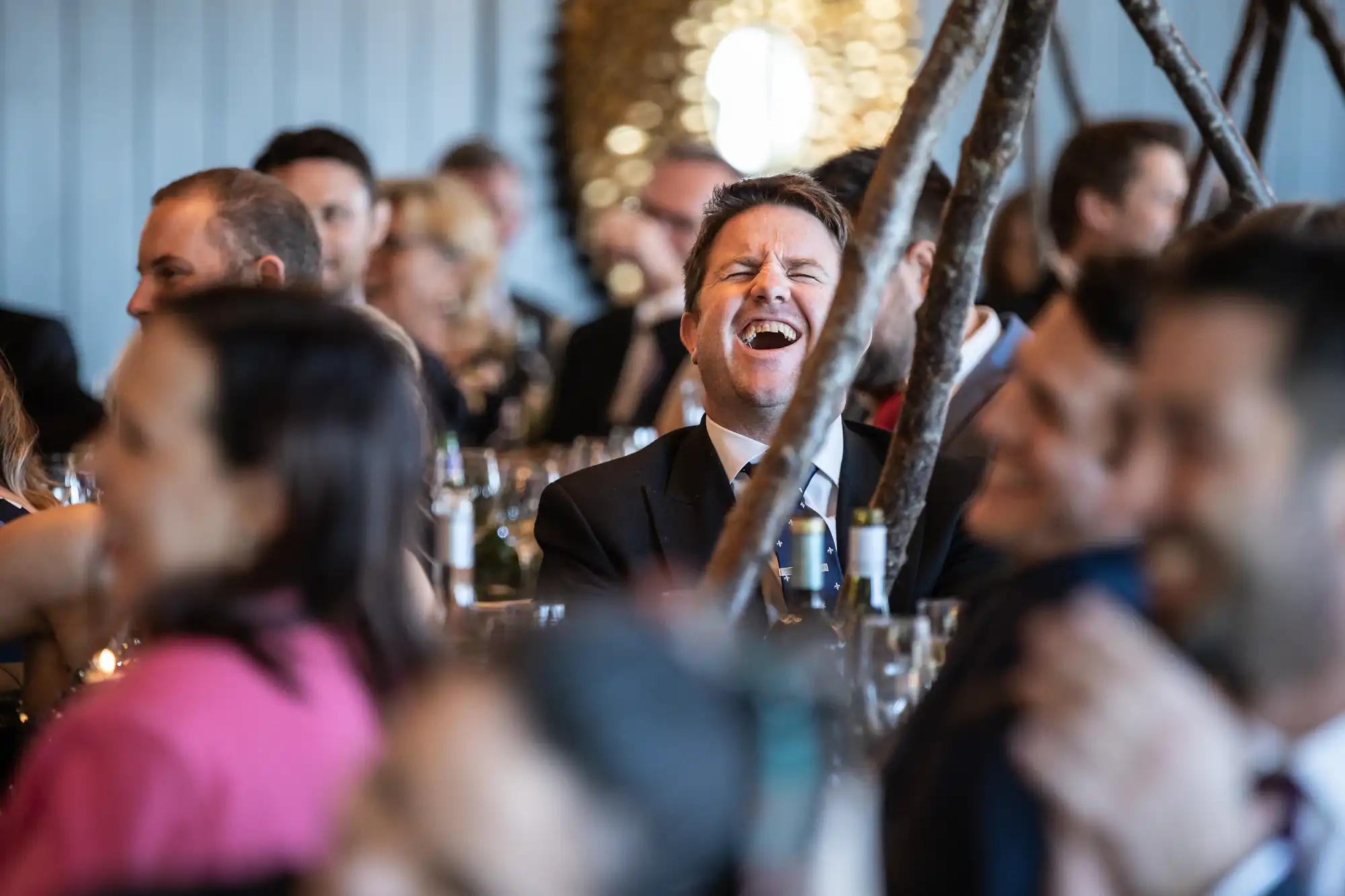 A man in a suit is laughing heartily while seated among a group of people at an indoor event.