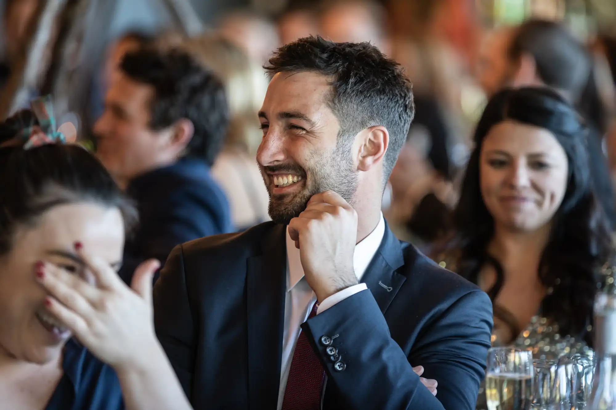 A man in a suit smiles and rests his chin on his hand while seated in a crowded room with other people.