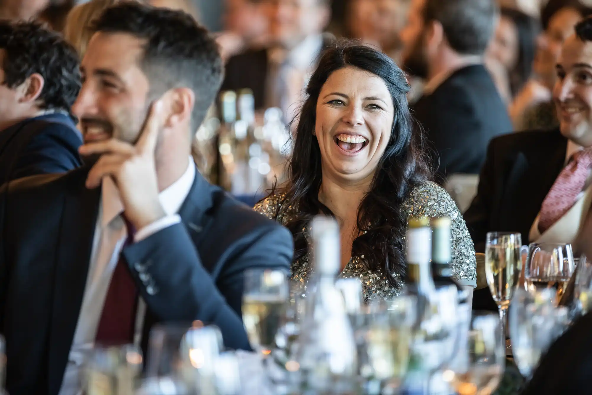 A woman in formal attire, smiling and laughing, is seated at a table with wine bottles and glassware, surrounded by other people in a lively event setting.