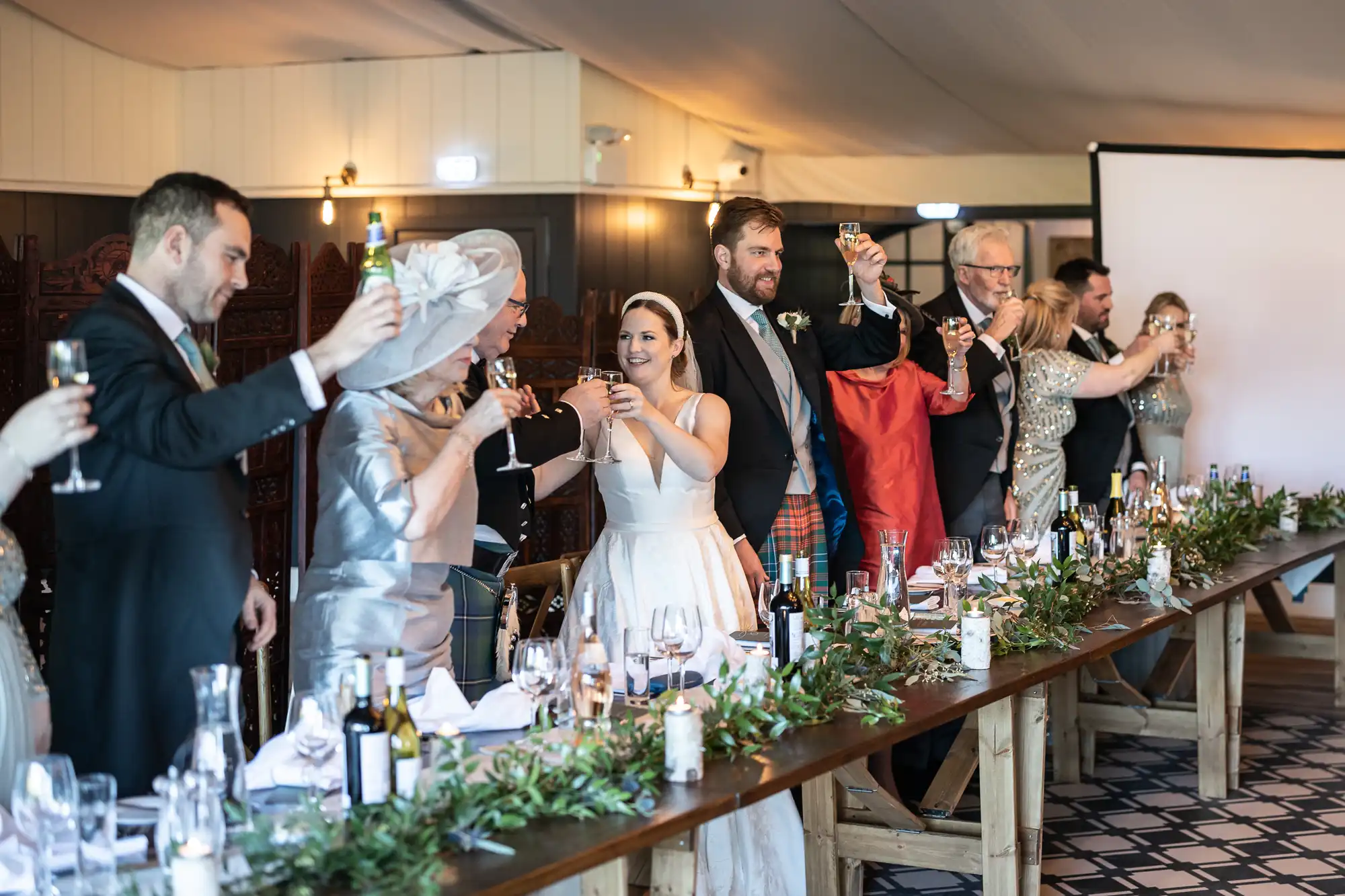 A wedding reception scene with a bride, groom, and guests raising their glasses for a toast while standing at a long, decorated table.