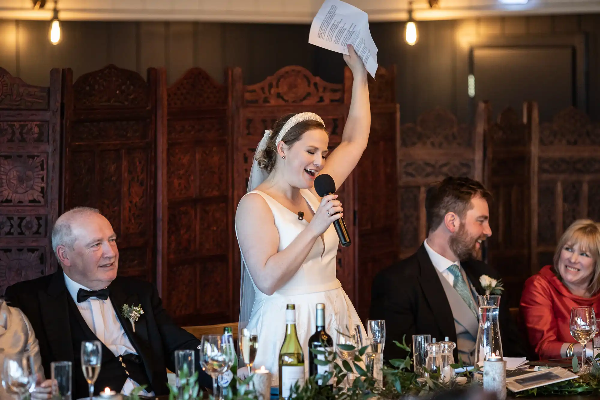 A woman in a white dress speaks into a microphone and holds up a paper, while sitting at a table with three other people in formal attire. The table is set with bottles, glasses, and decorative greenery.