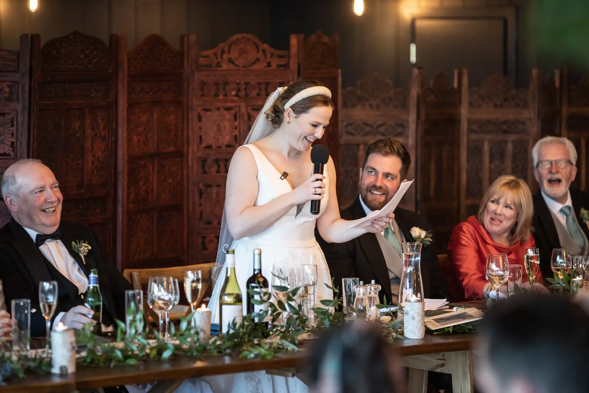 A bride stands and speaks into a microphone at a table during a wedding reception. Guests seated on either side of her are smiling and laughing. The table is decorated with greenery and glassware.