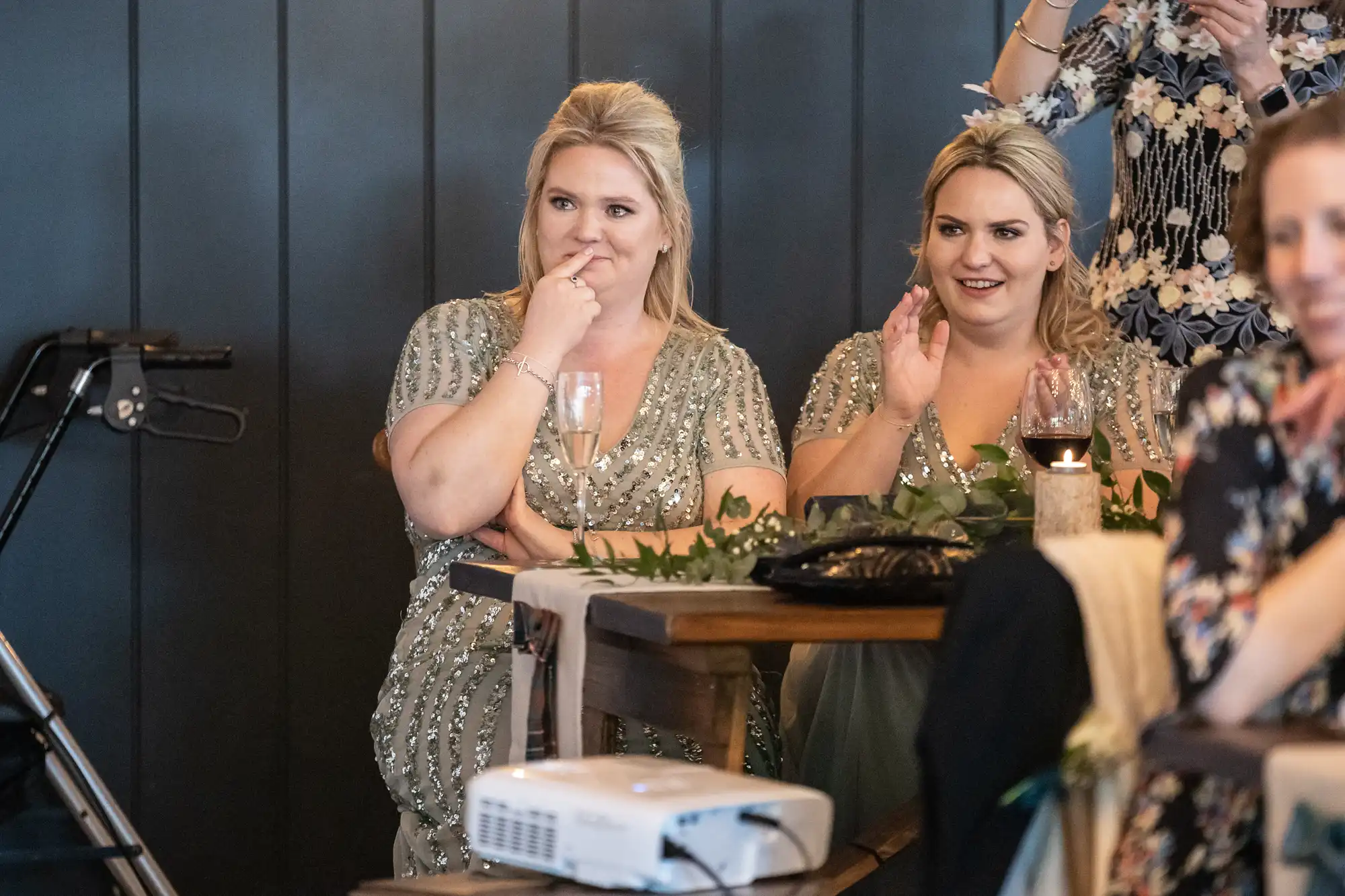 Two women in sparkly dresses are seated at a table, one holding a glass and smiling while the other claps. An overhead projector and greenery decor are visible in the foreground.